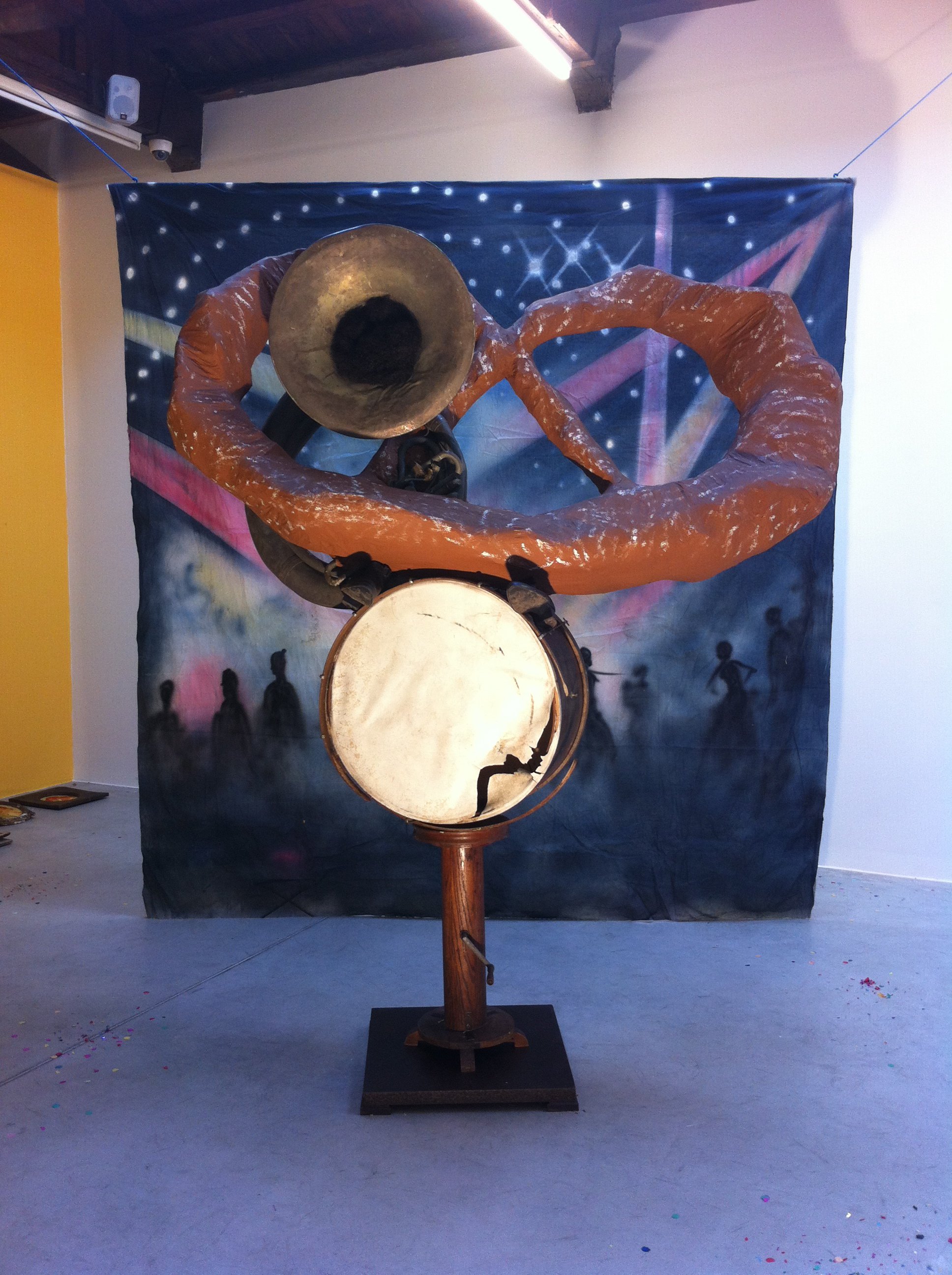 A large, abstract sculpture of a person with a big trumpet, wrapped in orange material, standing in front of a superhero-themed backdrop.