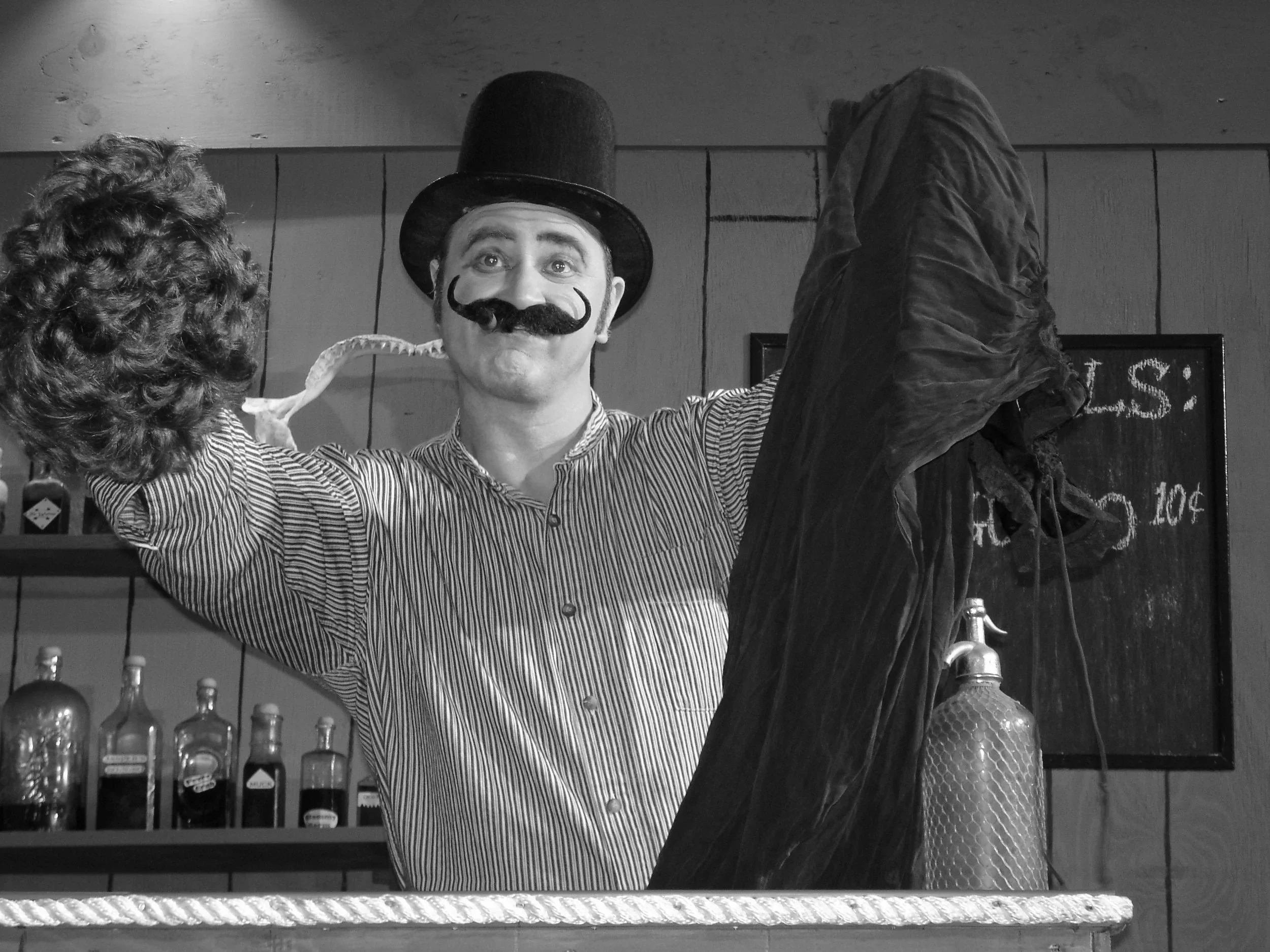 A man dressed as a vintage circus performer with a bowler hat, mustache, and striped shirt, holding a dark cloth or costume in a setting with bottles on a shelf and a chalkboard in the background.