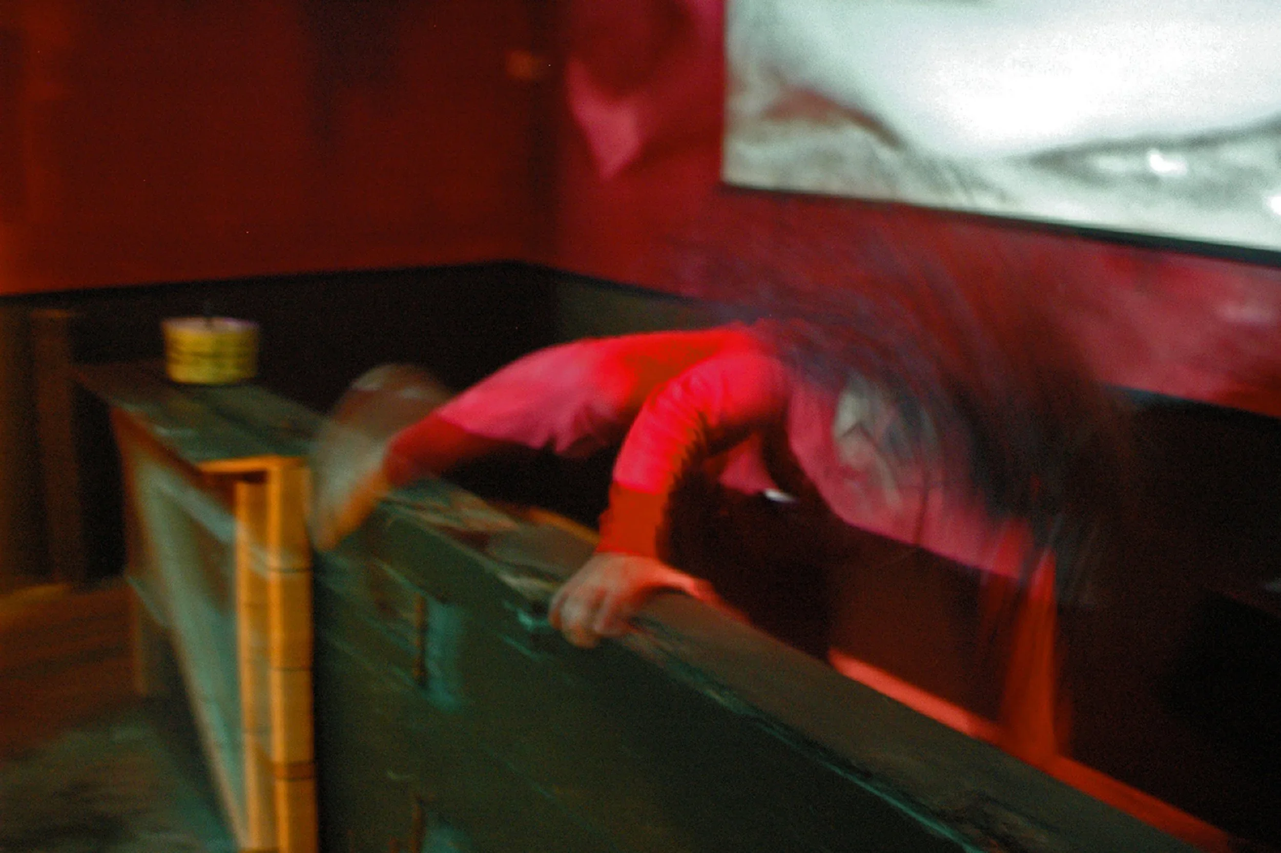 A person with long dark hair leaning over a black table or counter, wearing a red and white jacket, in a dimly lit room with red and black decor.