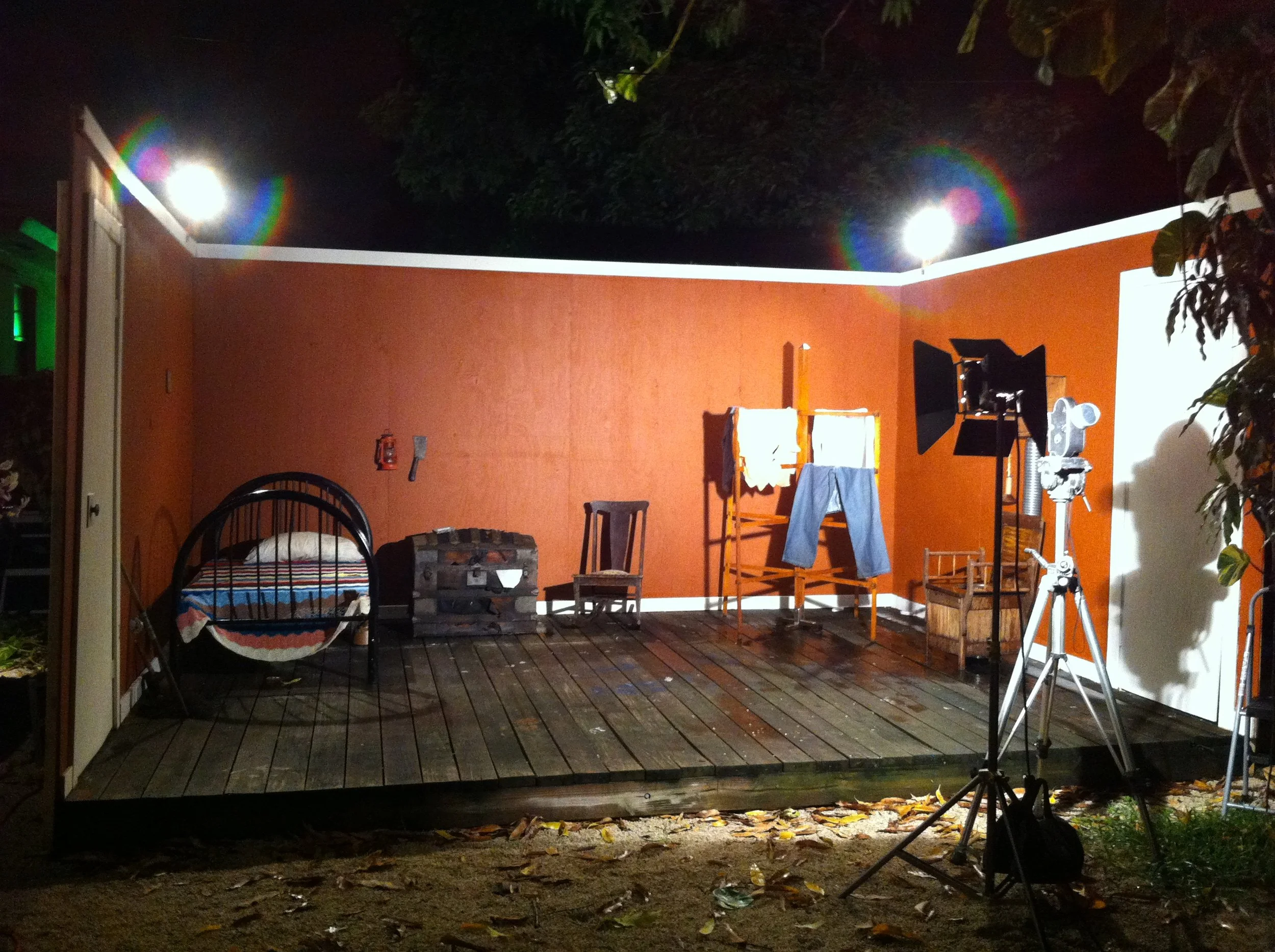 Outdoor stage with orange wall backdrop, set up with vintage furniture, clothes drying rack, professional lighting, and camera equipment under night sky with bright lights and rainbow flares.