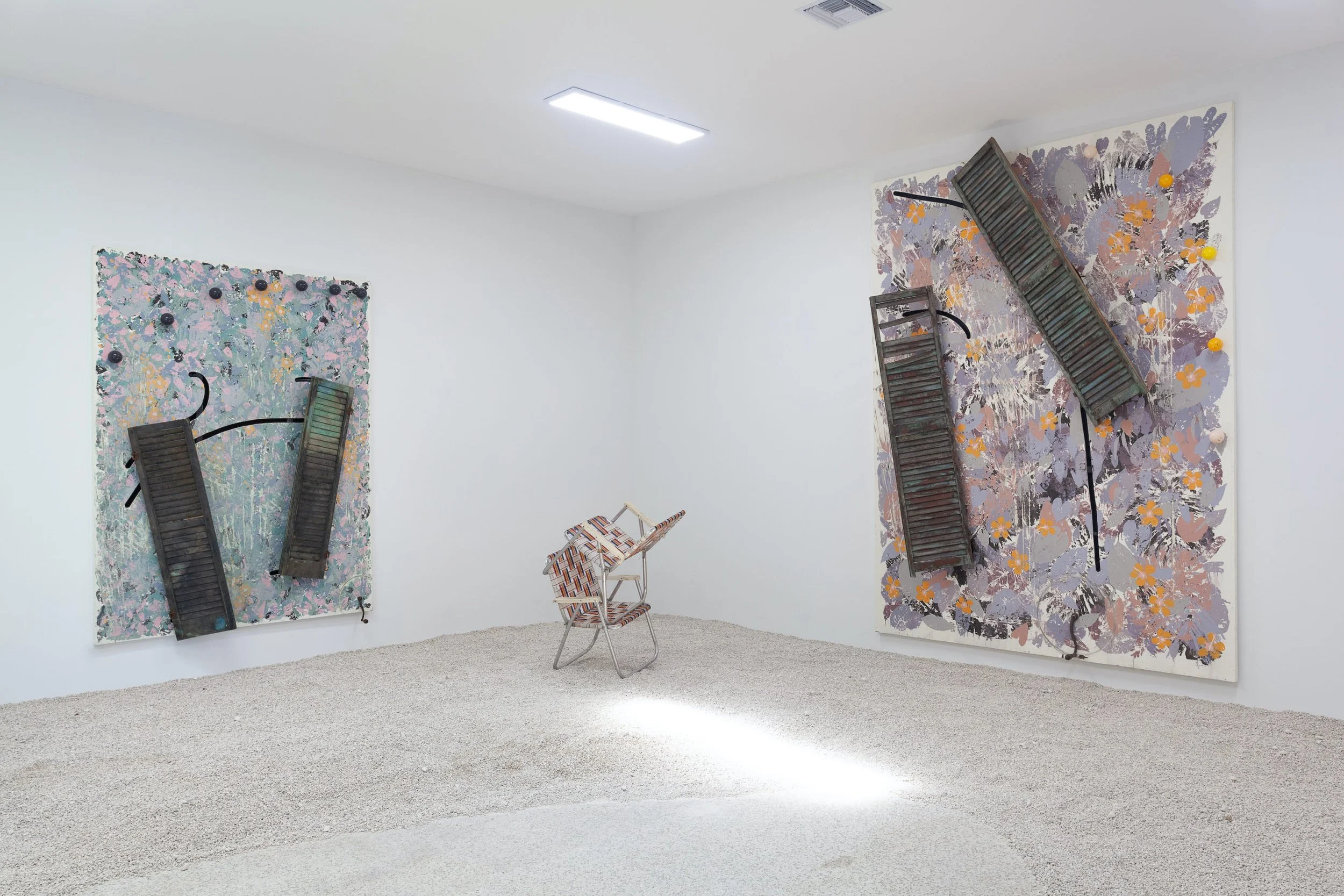Art installation featuring two large canvas artworks with wooden shutters attached, a small patterned chair, and a plain white room.