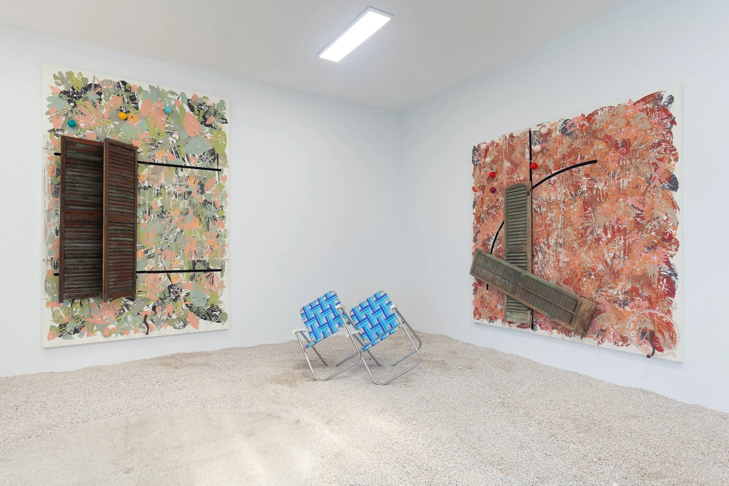 Contemporary art exhibit featuring two large mixed media canvases with wooden shutters and colorful abstract elements, and two blue patterned folding chairs on a light gravel floor.
