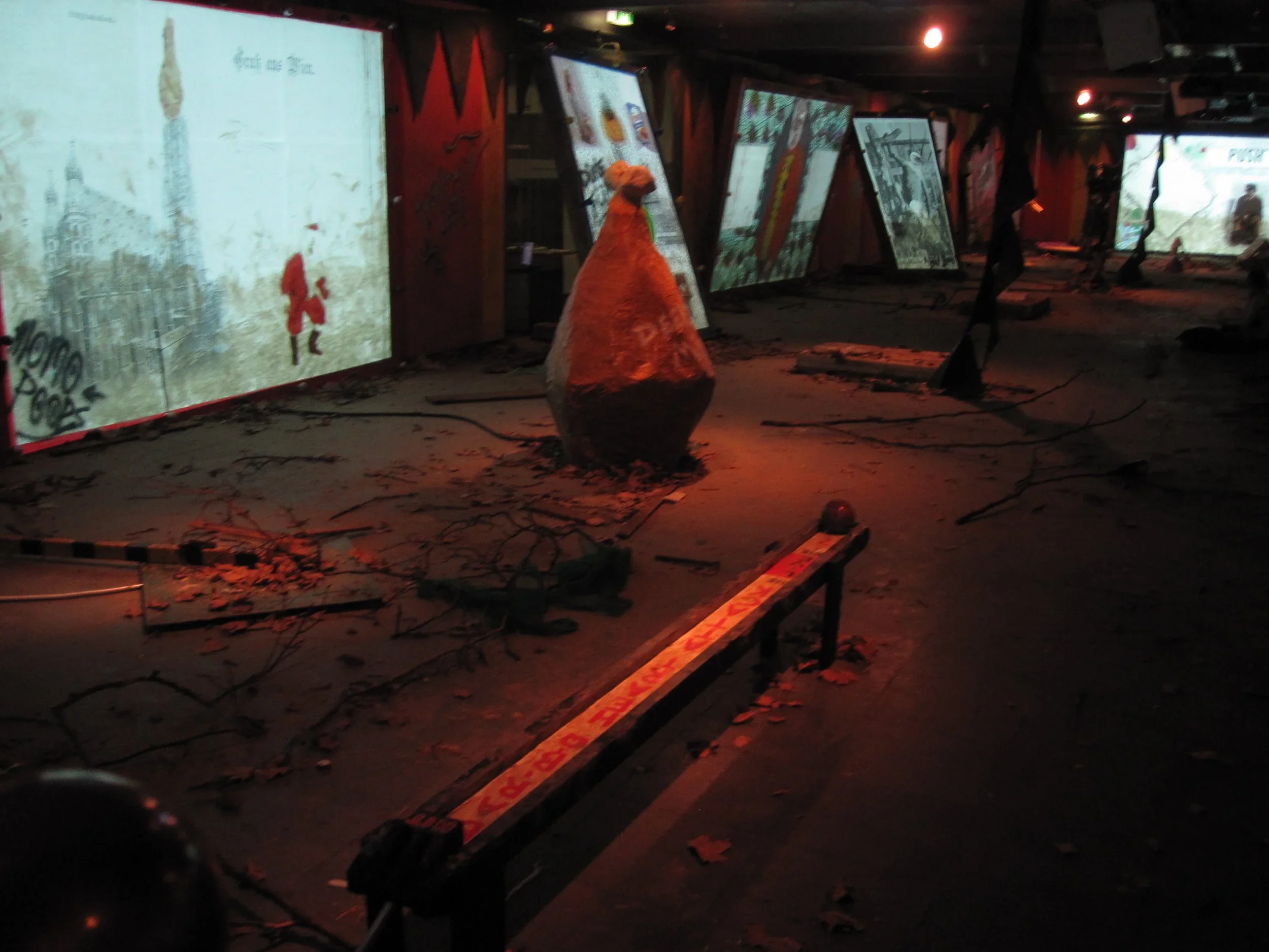 Dark indoor space with multiple screens displaying images, debris on the ground, and a large cone-shaped object in the center. The screens appear damaged or part of an exhibit.