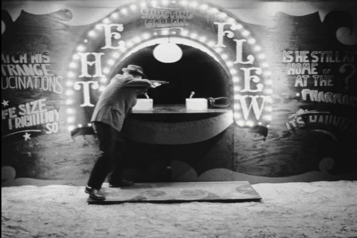A man standing on a step stool aiming a gun at a shooting game at a carnival or arcade. The background features a round illuminated sign with the word 'THE FEELING' and some painted text about the game.