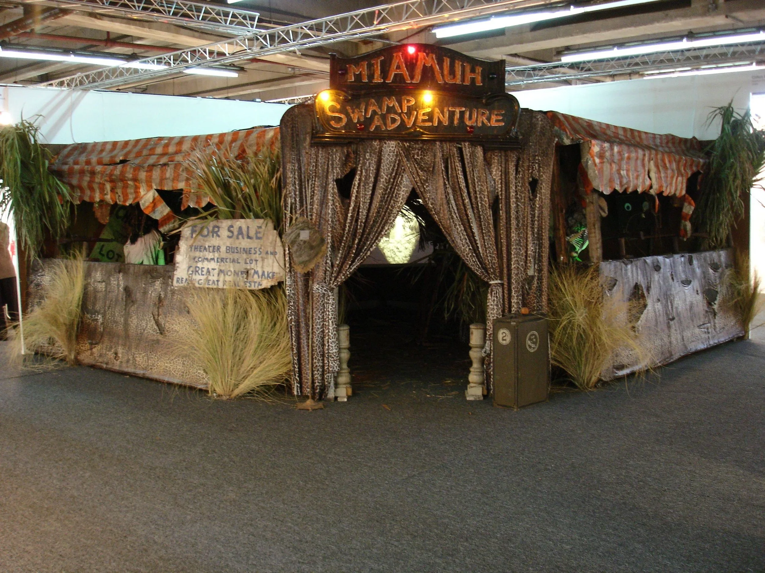 Entrance to a themed attraction called 'Miami Swamp Adventure' with a jungle style decor, camouflage curtains, plants, and a sign indicating the attraction is for sale.