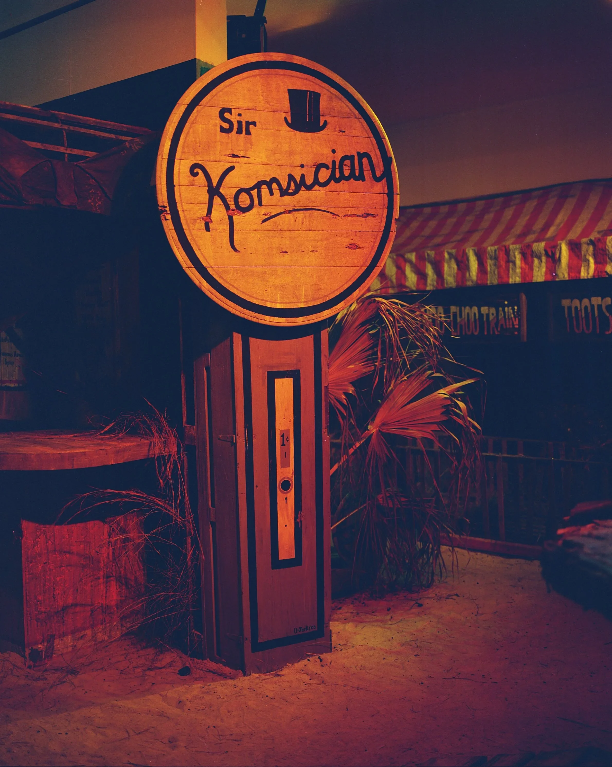 A wooden sign with a top hat icon and the words "Sir Komsician" on a beach, with sand, plants, and a striped canopy in the background at night.