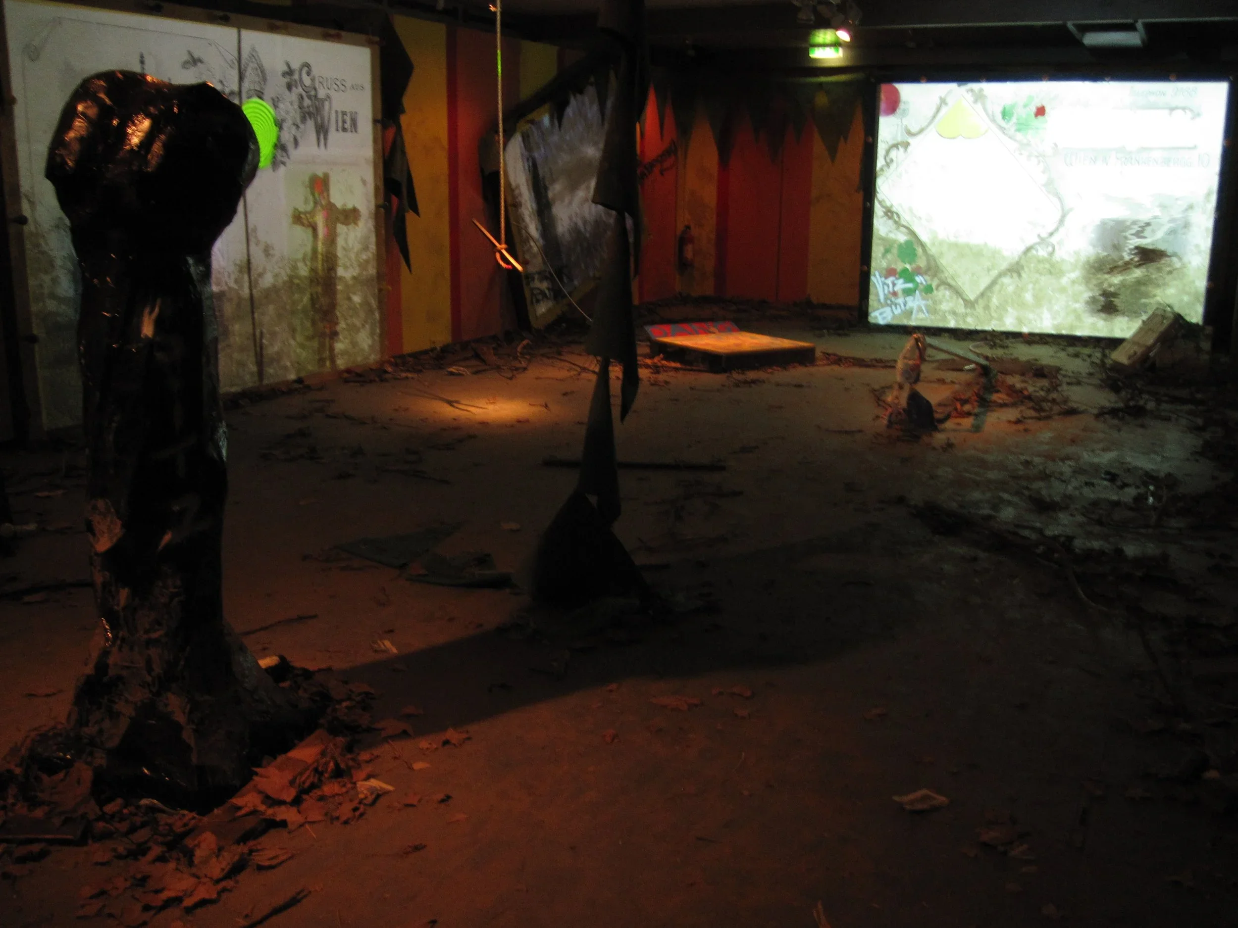 The image shows an indoor scene with dim lighting, debris scattered on the floor, and a large screen or projection in the background displaying a map or landscape. The space appears to be abandoned or damaged, with dark-colored flags or banners hanging from the ceiling and various damaged objects around.