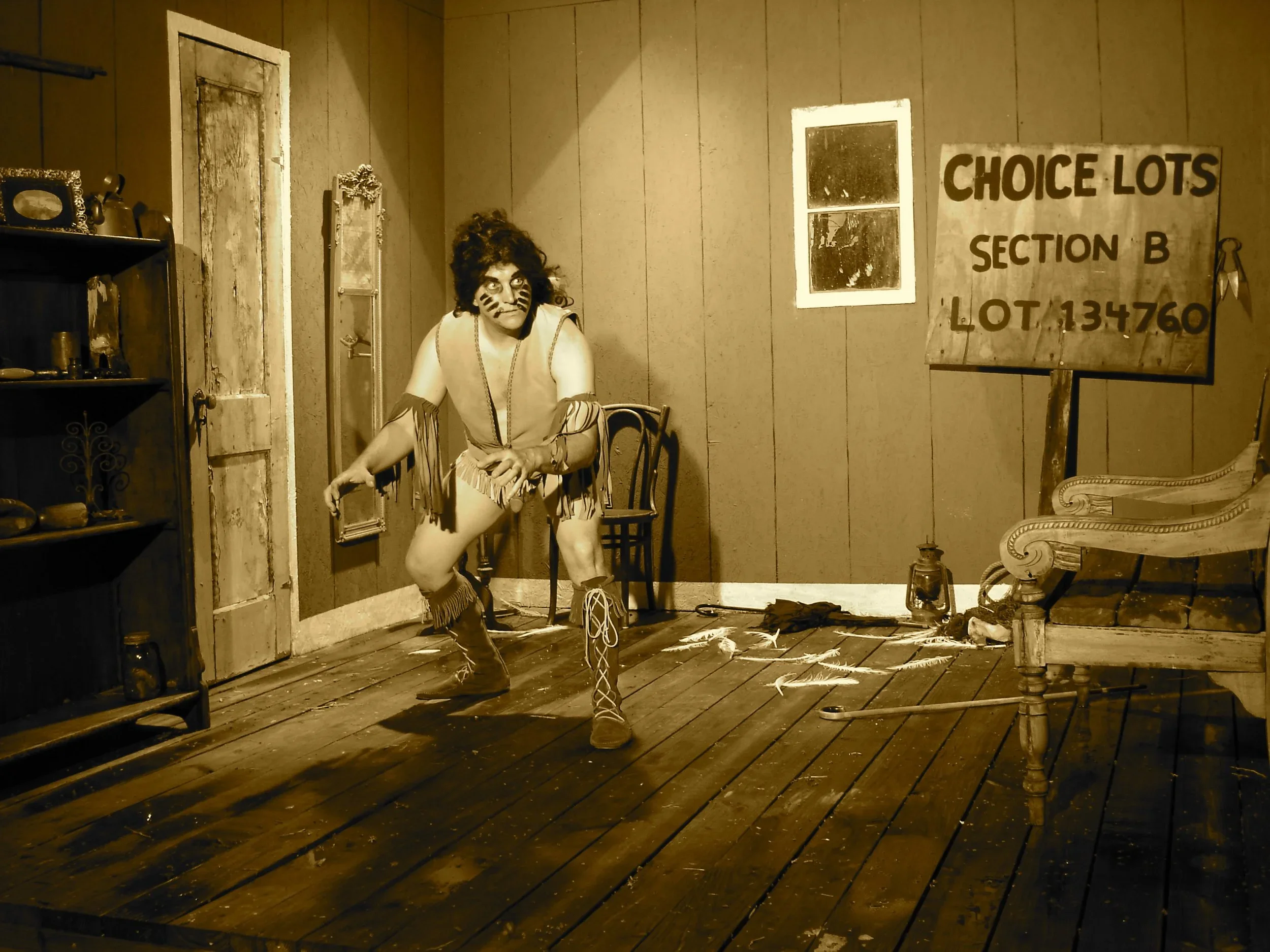 A person dressed in a Native American costume with face paint, standing on a wooden stage in a room with wood-paneled walls. There is a sign that reads 'Choice Lots Section B Lot 134760' on the wall. The room contains a wooden shelf with objects, a chair, a bench, and a small window.