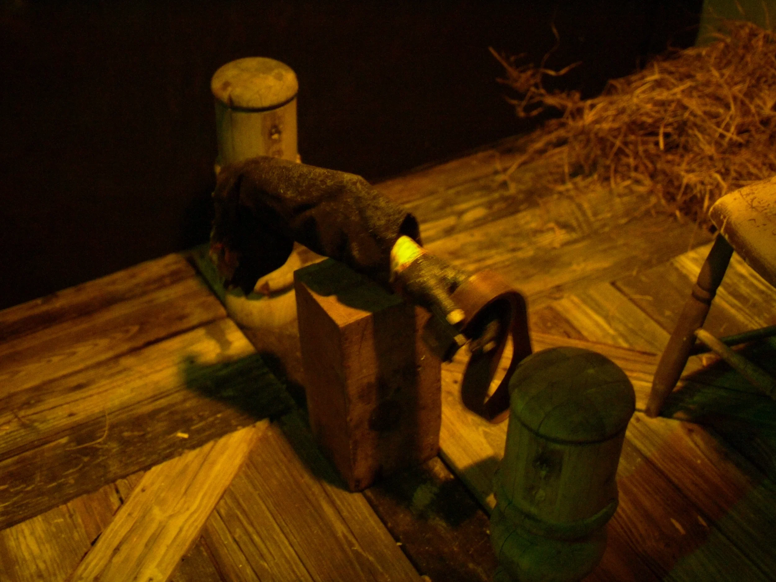 A stuffed raccoon toy lying on a wooden dock at night, with a dock post and a chair visible nearby, and some straw or dried grass in the background.