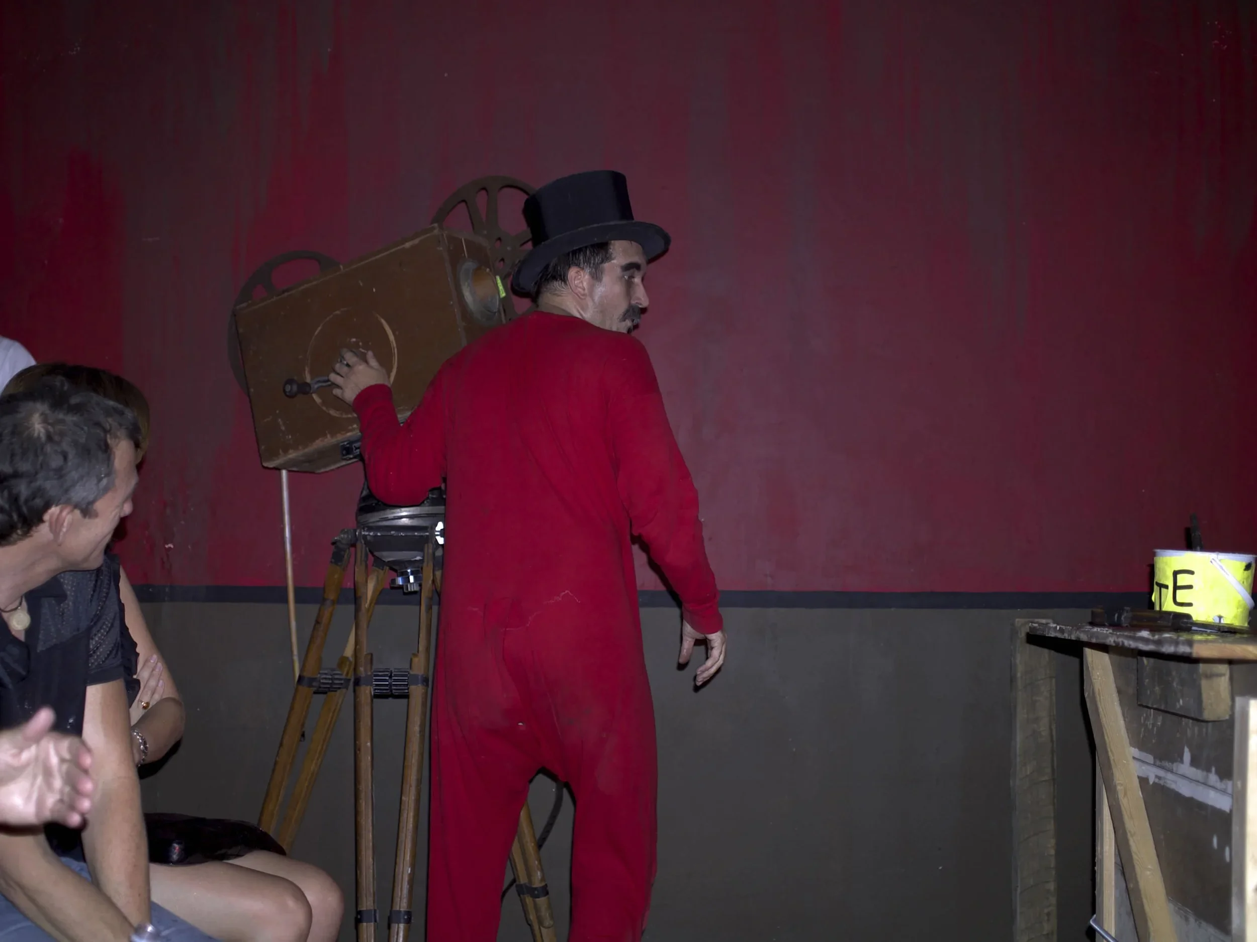 A performer dressed in a red costume and black top hat stands in front of a vintage movie projector on a tripod, with a red wall backdrop and several audience members watching.