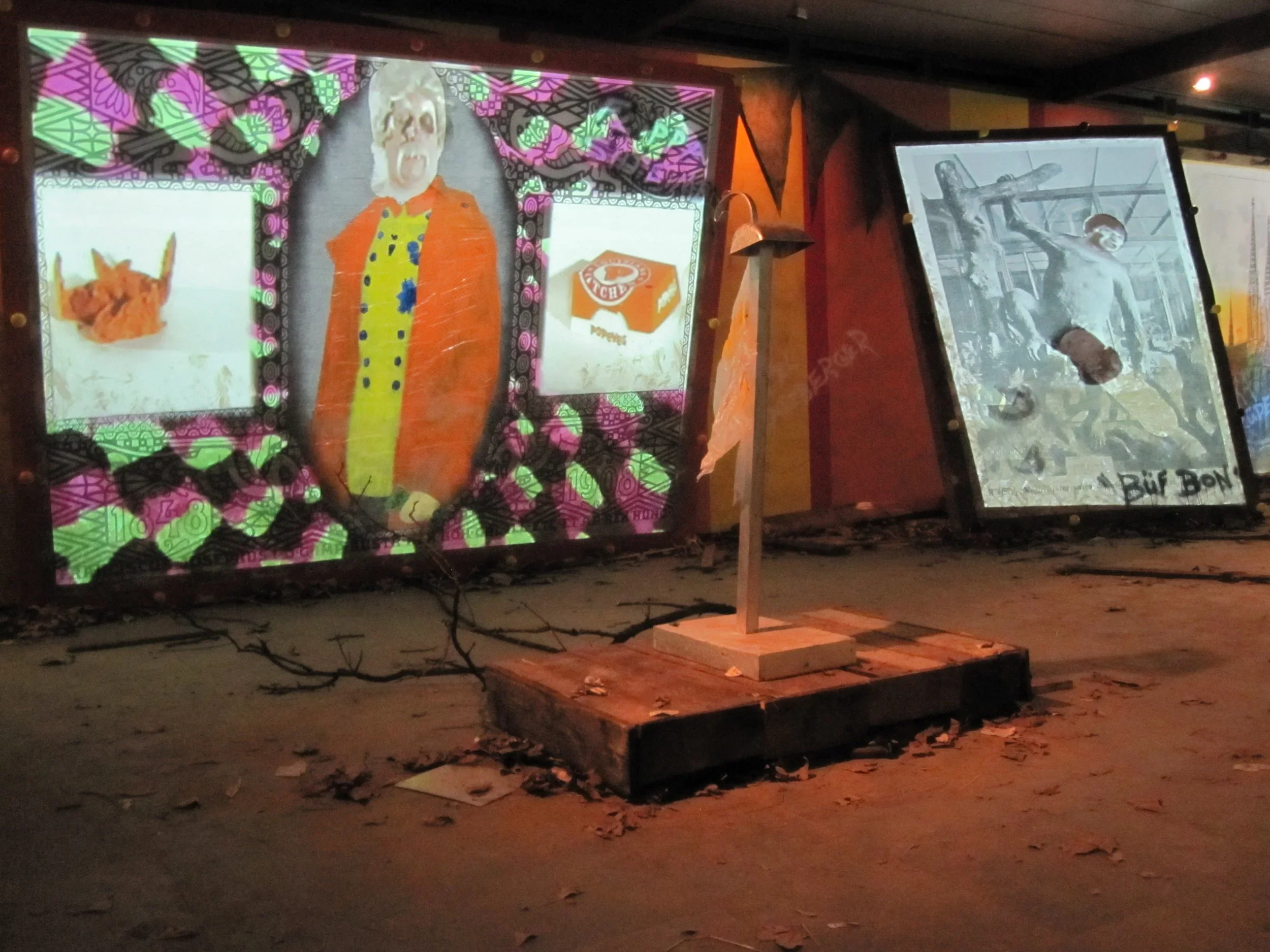 Two illuminated art pieces displayed outdoors at night on a sidewalk. The left art features a colorful, cartoon-like figure with a large, white face and orange hair, surrounded by bright patterns and two smaller images of food items. The right art is a black and white drawing of a man with a muscular build, holding a sword, with text reading 'BUFF BOND' at the bottom.