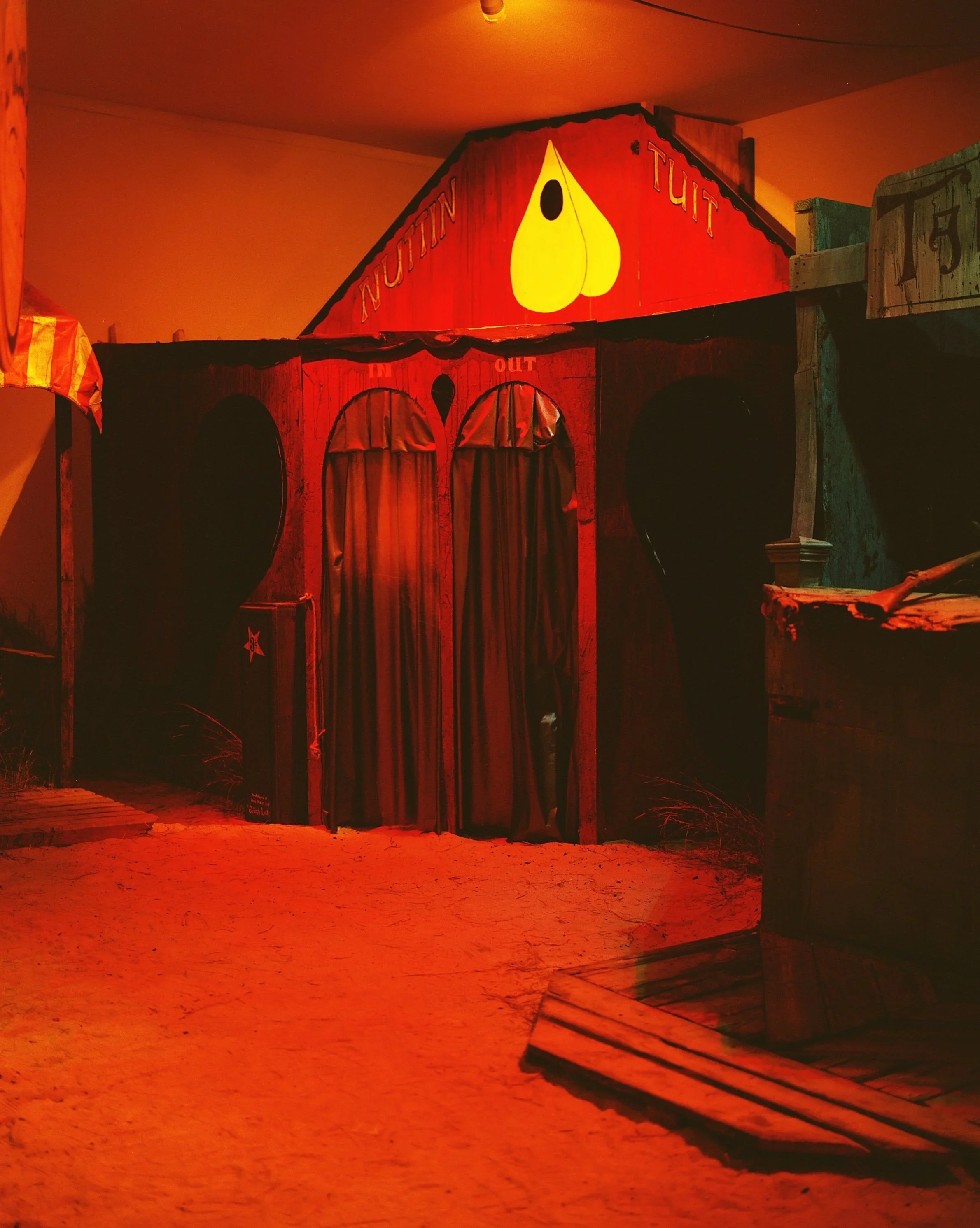 A spooky haunted house-themed attraction with wooden walls painted red, featuring a sign with a yellow teardrop shape and the words "Nut" and "Tuit". The house has three arched doorways covered with curtains, and a glowing orange-red lighting creates an eerie ambiance.