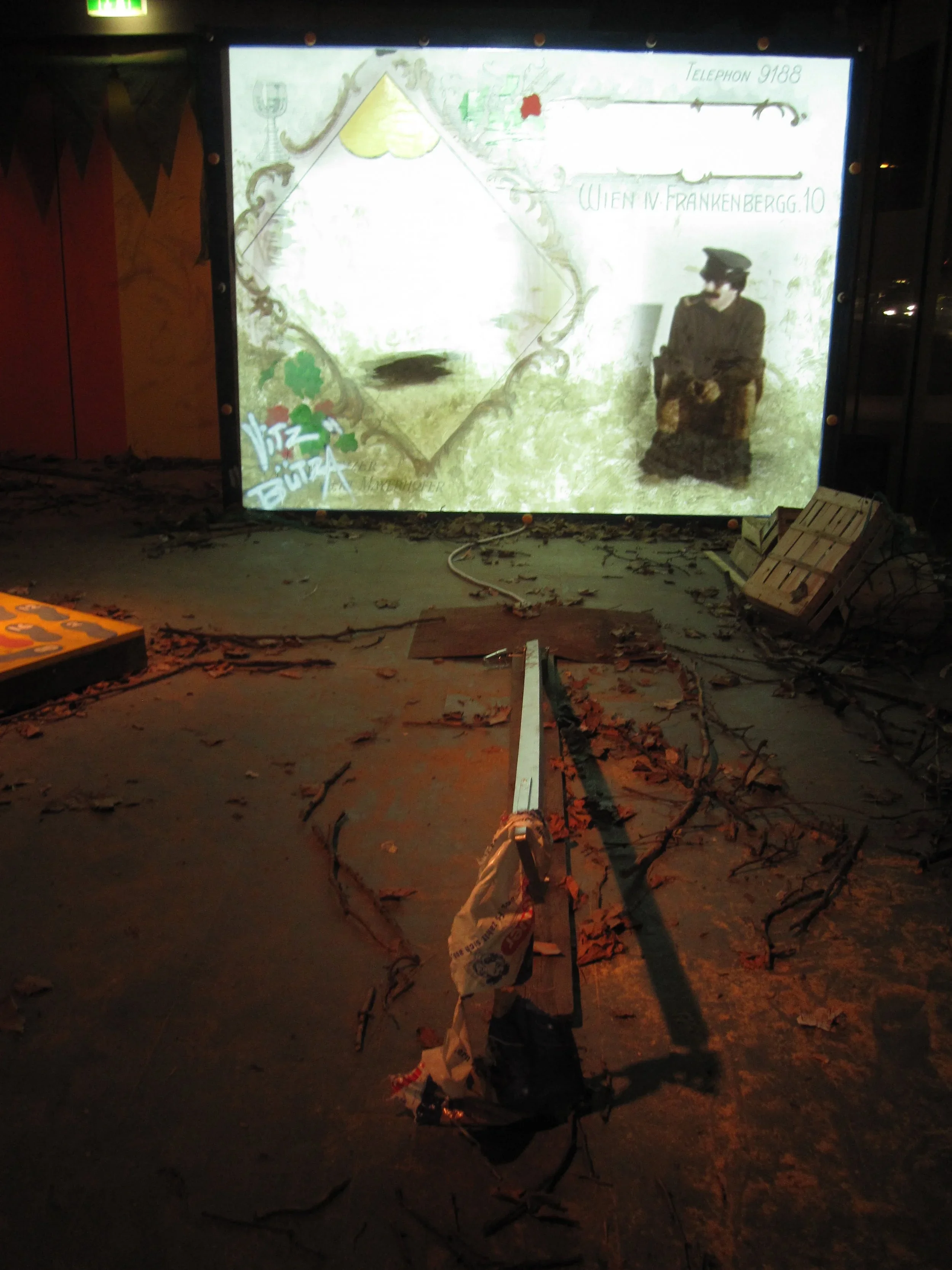 Photograph of a damaged projection screen with graffiti and an old photograph of a woman in a dress and hat, in a dimly lit room with a fallen light fixture and scattered debris on the floor.