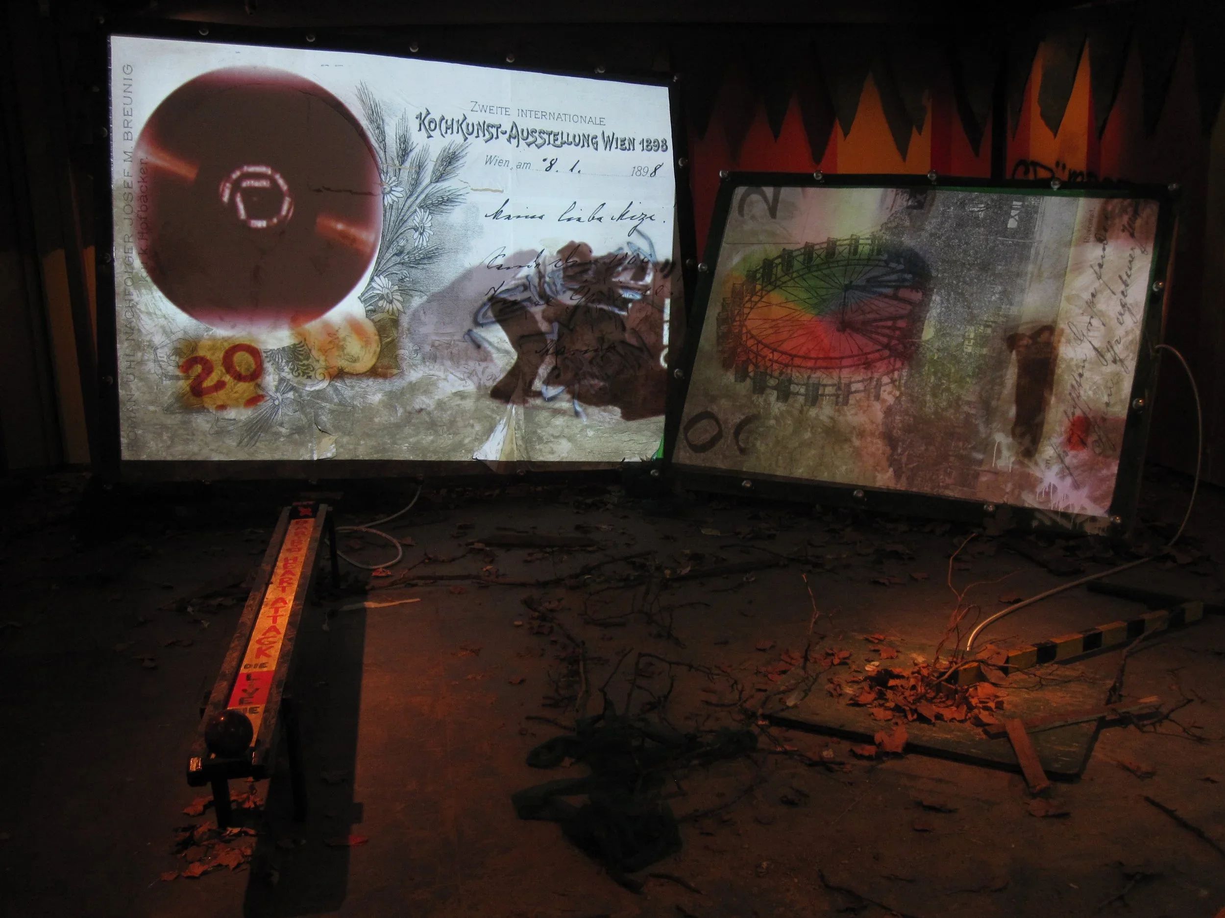 Two digital screens display images related to art exhibition, one featuring a vintage postcard and a large colored ball, the other showing an abstract image of a carnival Ferris wheel, with a floor scattered with fallen leaves and a caution barrier in the foreground.