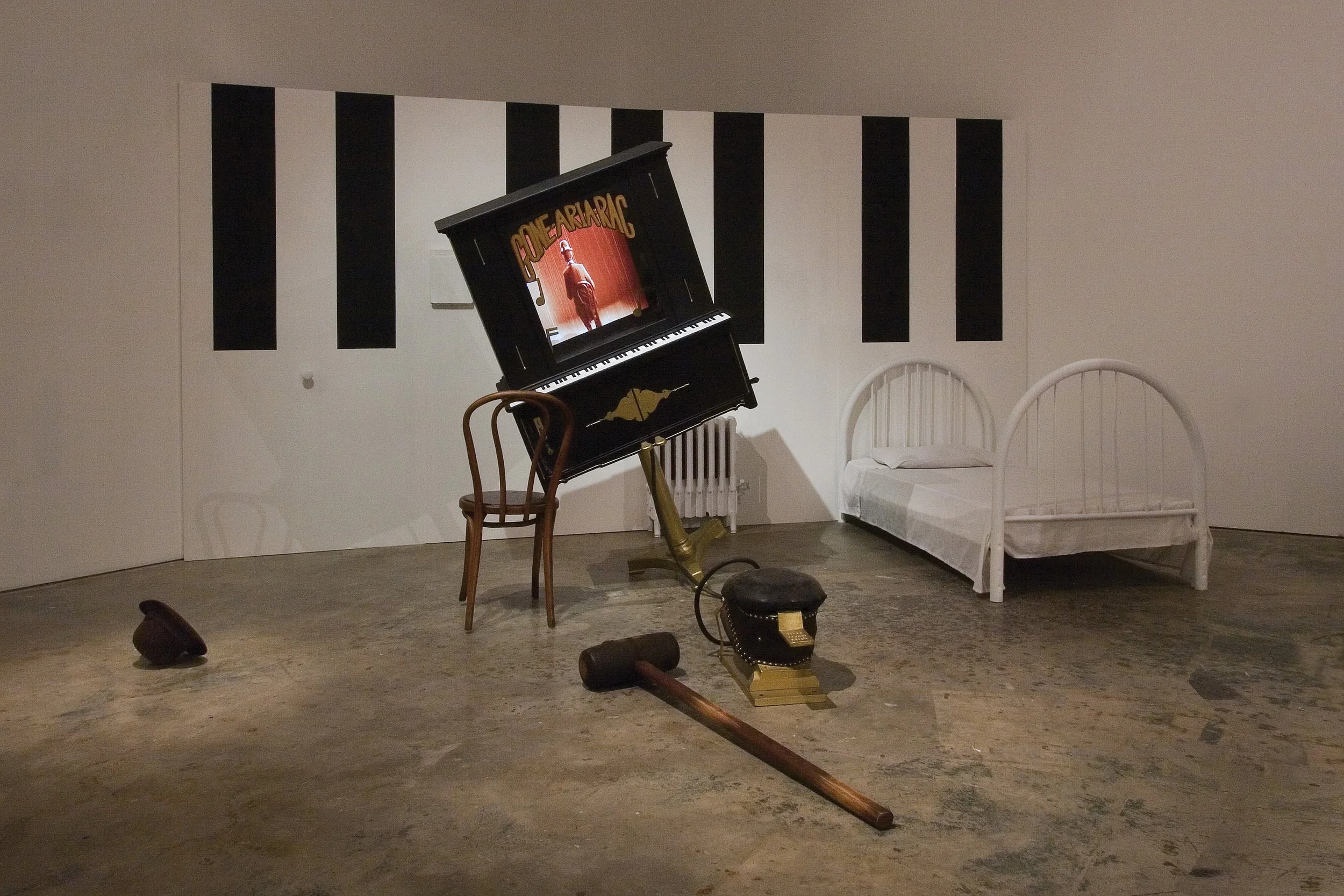 An art installation featuring a painted black upright piano with the words "Bone Ar ARAC" on its lid, a wooden chair, a small black stool, a long wooden mallet on the floor, a white bed with a curved headboard, and a wall mural with black vertical stripes.