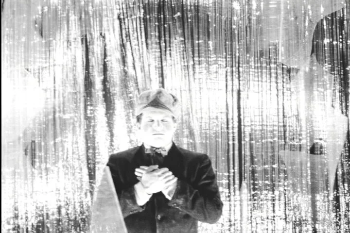 A person standing in front of a curtain with a shiny, textured background, wearing a hat and coat, holding their hands over their chest.