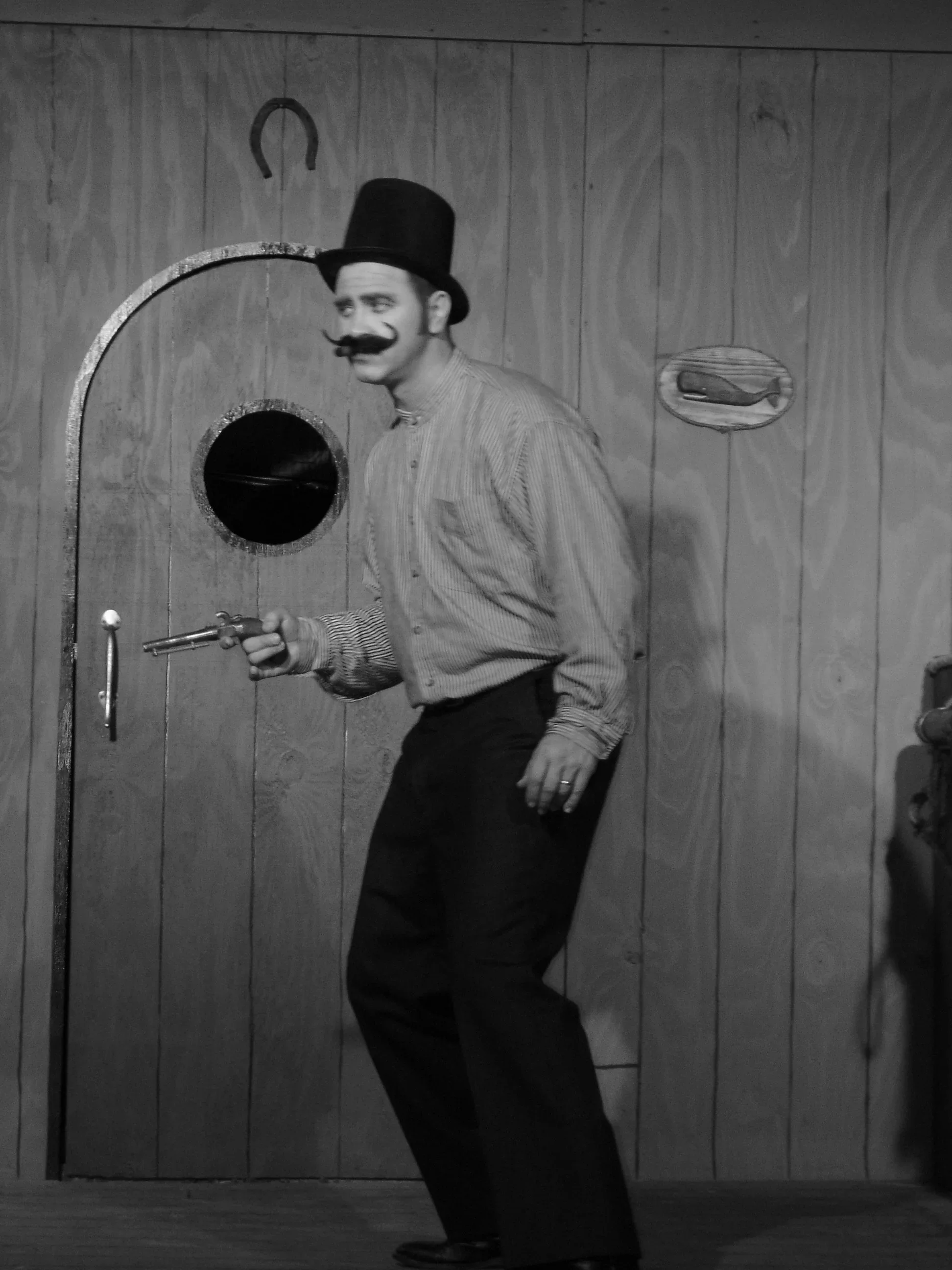 A man dressed as a cowboy or western character with a mustache, top hat, and striped shirt, holding a toy gun, standing in front of a wooden wall with a horseshoe and a small sign.