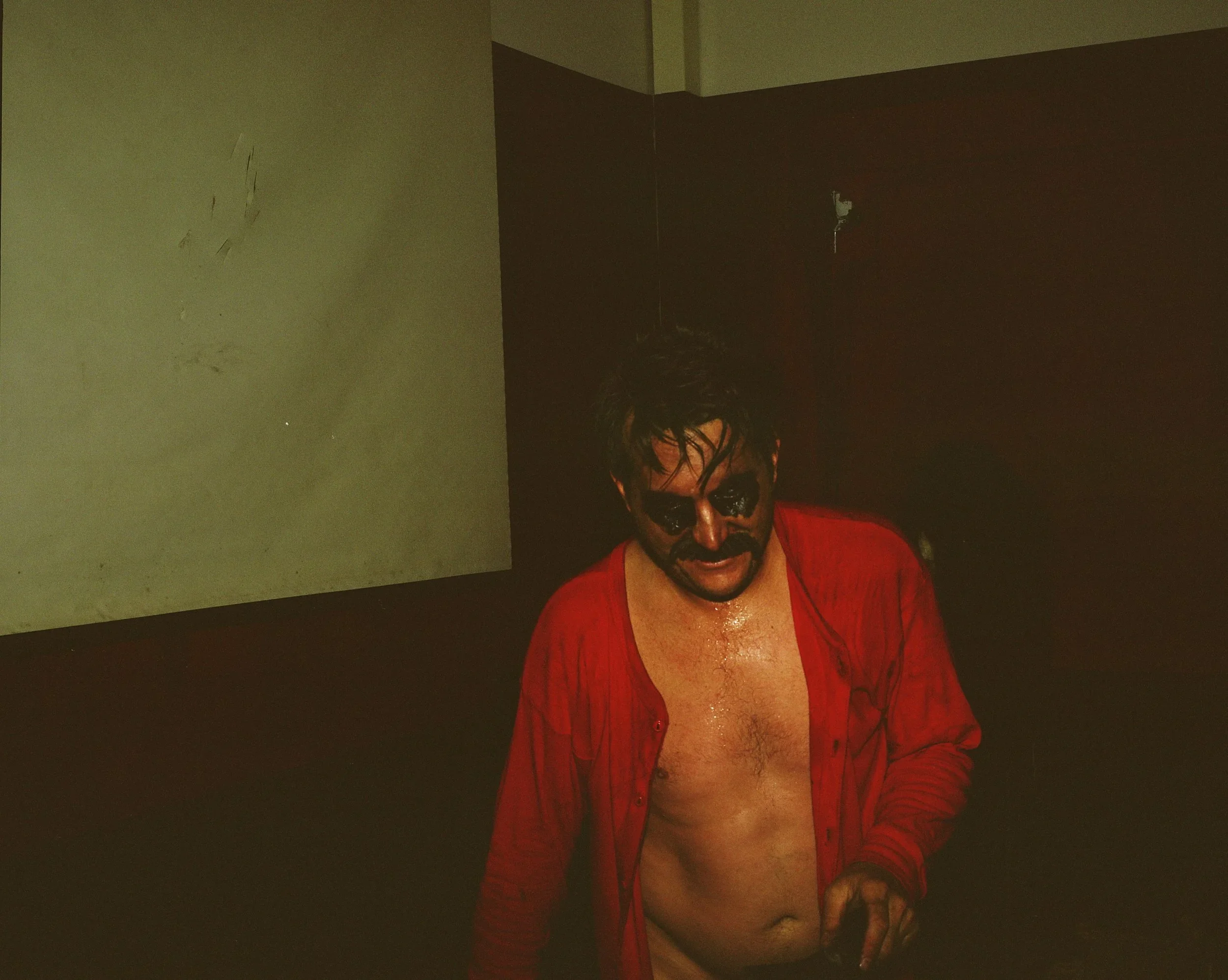A man with black face paint and messy hair, wearing a red button-up shirt open to reveal his chest, is standing in a dimly lit room with black and gray walls, appearing sweaty or wet.