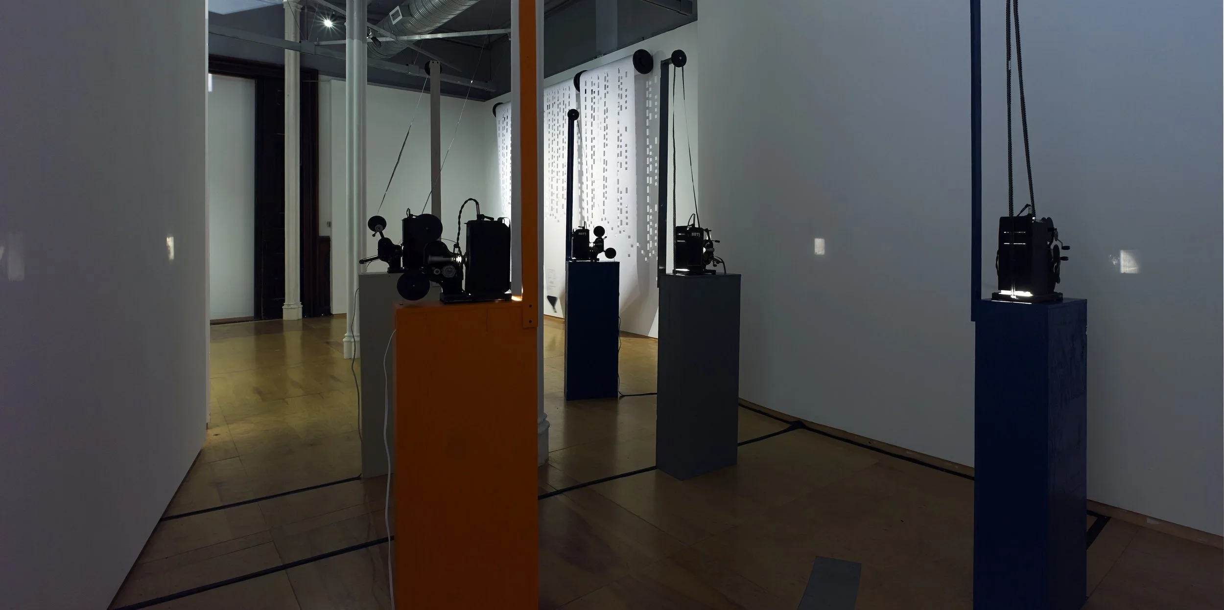 Contemporary art installation featuring black mechanical devices mounted on colorful pedestals in an art gallery with wooden floors and white walls.