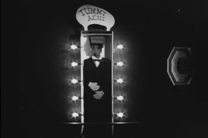 A person in a tuxedo inside a photo booth with a marquee light frame. The booth has a sign that says 'TUMMY ACHE' at the top, and the person is wearing a bow tie.