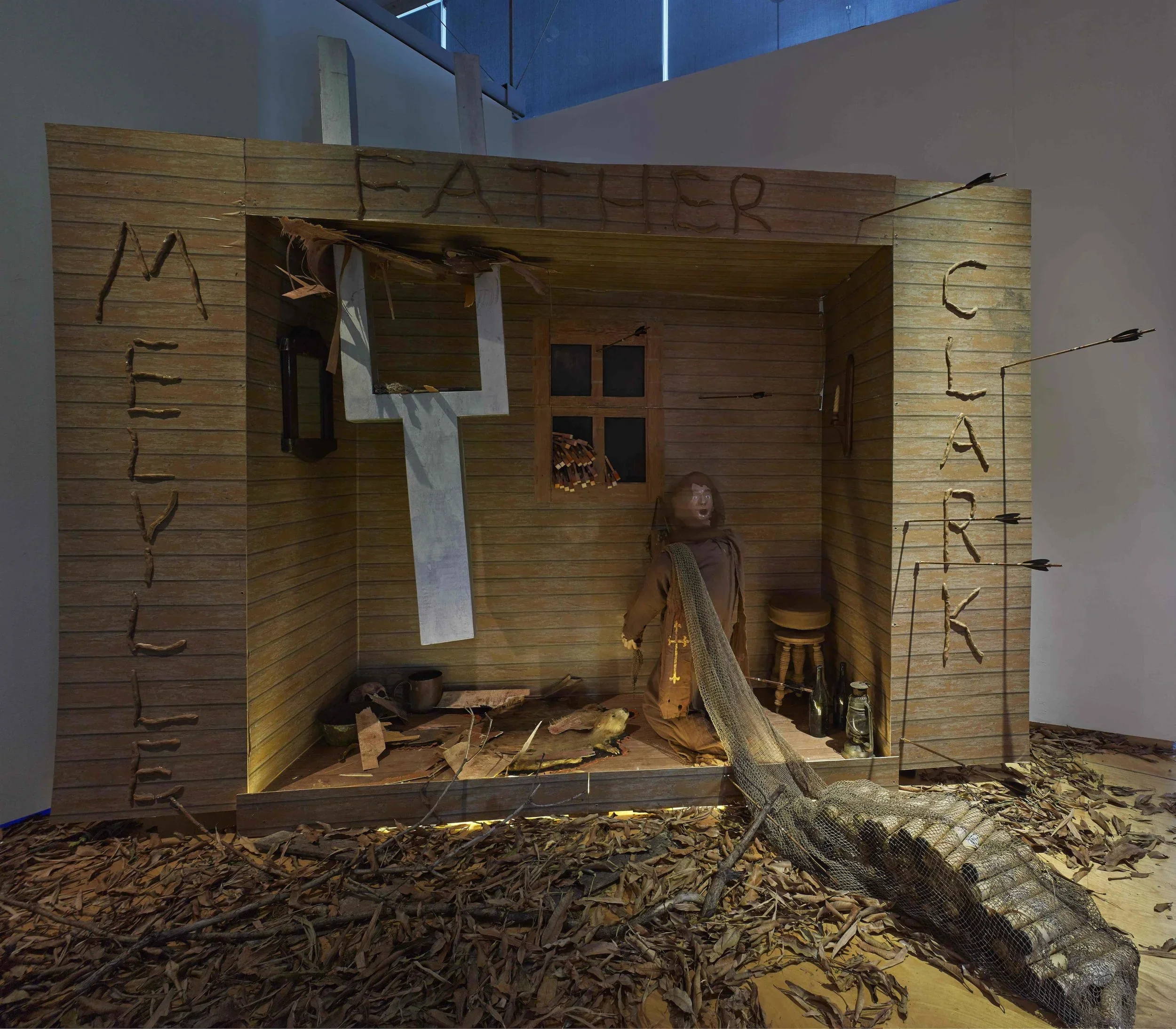 A wooden art installation depicting a historical scene with a kneeling figure surrounded by leaves and objects, with the words 'MEET THE FIGURE', 'FATHER', and 'TEACHER' carved into the wooden frame.