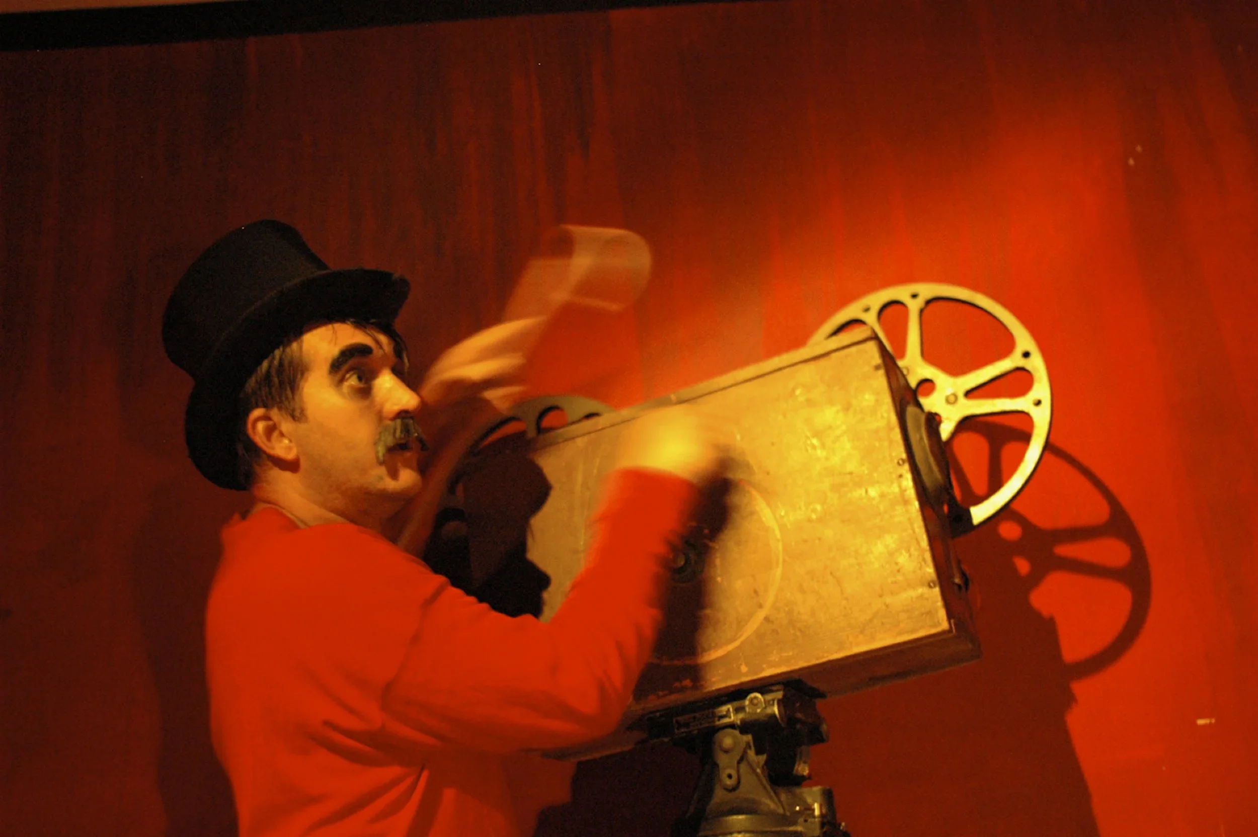 A man dressed as a vintage showman or performer, wearing a top hat and red costume, operating an old film projector against a red background, with a shadow cast on the wall.