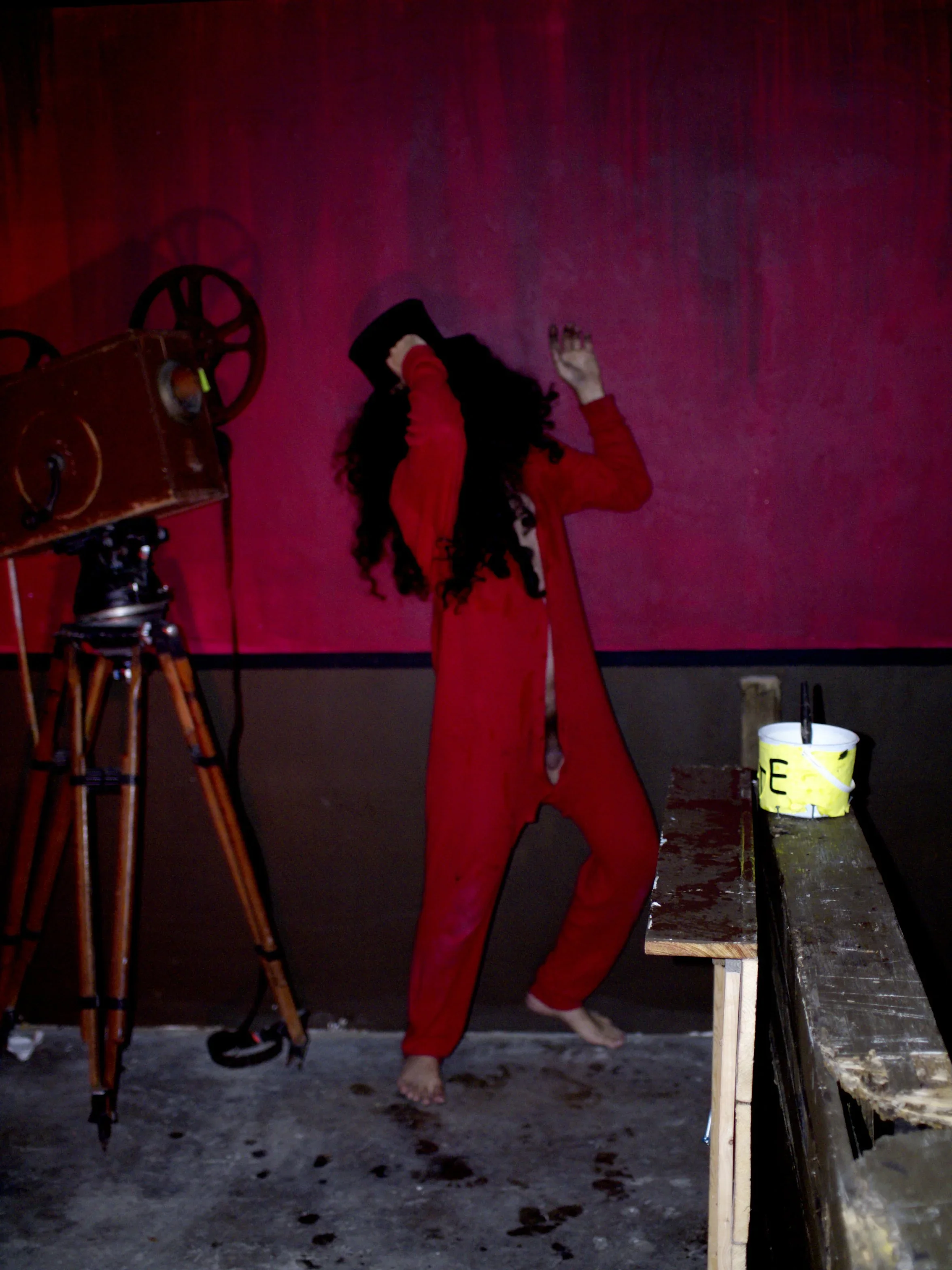 Person with long curly hair in red pajamas, holding a black bucket upside down over head, standing on a dark, worn floor in front of a pinkish-red wall, with a vintage film camera and a yellow bucket on a wooden table nearby.