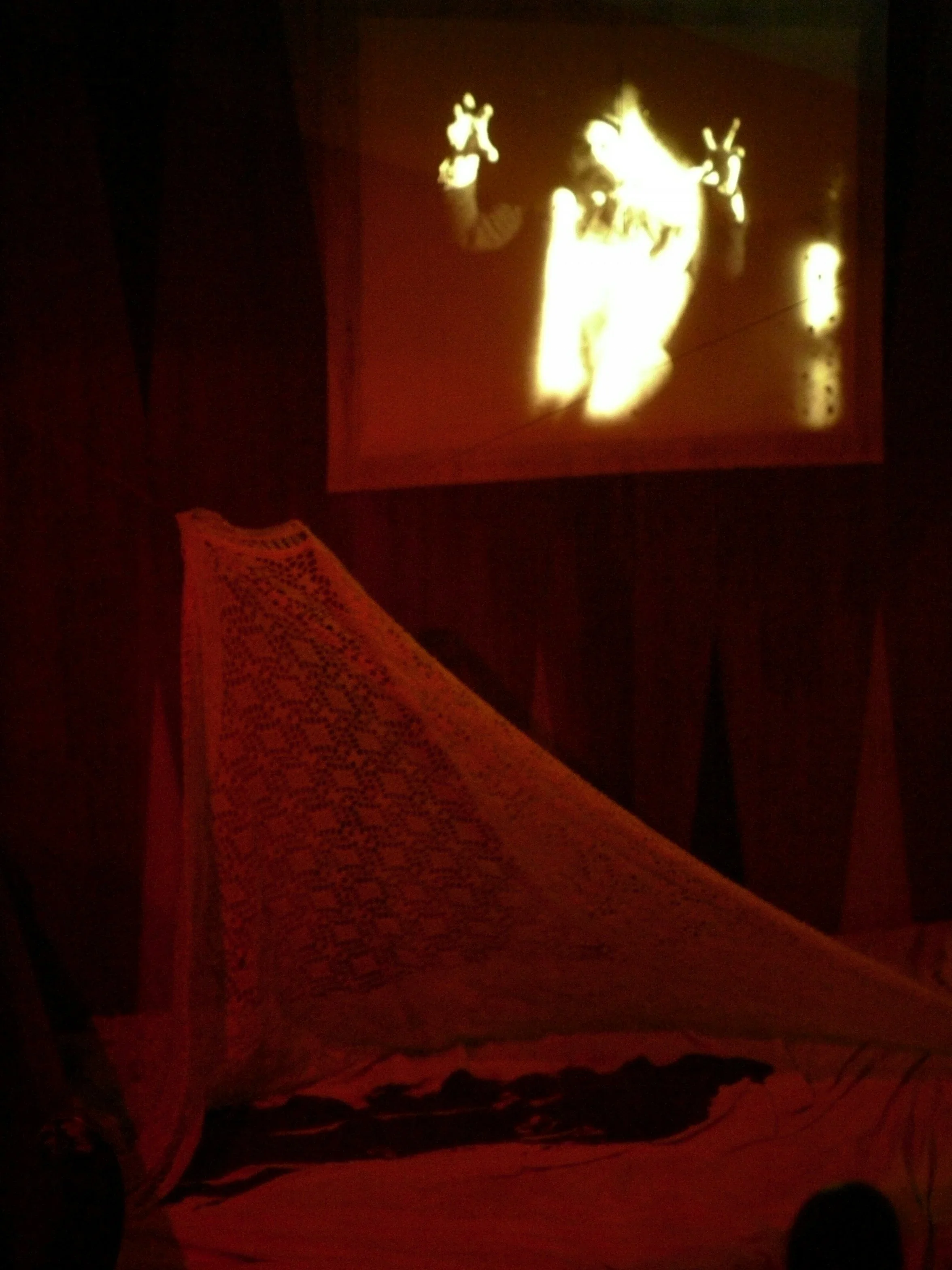 A shadow puppet show projected onto a wall, featuring a character with pointed ears and hands making peace signs, with a red curtain or backdrop.
