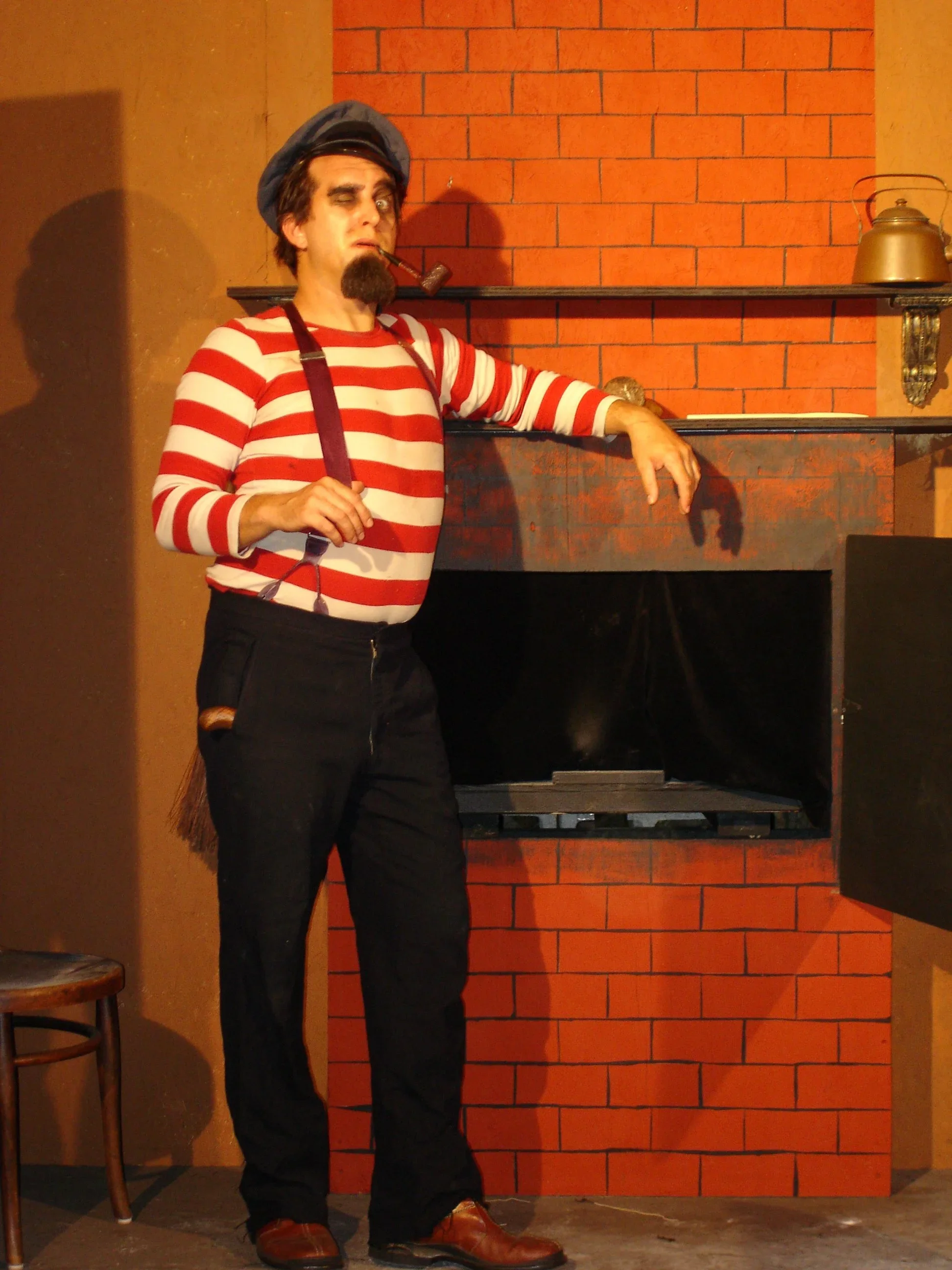 A man dressed as a mime in a striped red and white shirt, black pants, and a black cap, standing in front of a fireplace with a brick wall. He has a pipe in his mouth and is striking a theatrical pose.