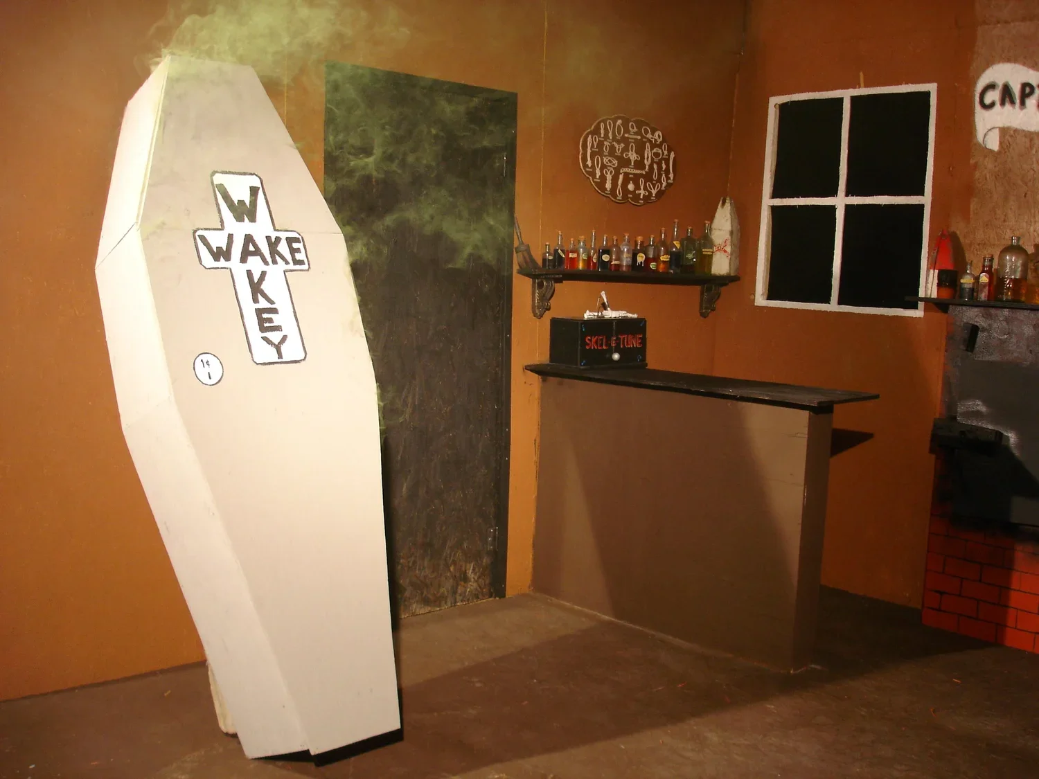 Bar scene with a white coffin-like object displaying the word "WAKKEY" with a cross-shaped design. The background has a brown wall, a shelf with bottles, a window with black panes, and a small bar counter creating a cozy, edgy atmosphere.
