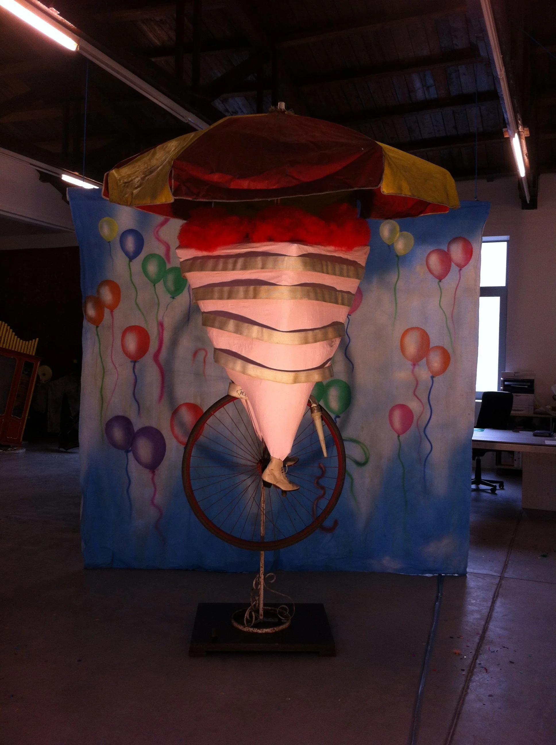 An artistic sculpture of a spinning top with a bicycle wheel frame, topped with a red and yellow umbrella and decorated with pink, beige, and green ribbons, set against a colorful balloon-themed backdrop.