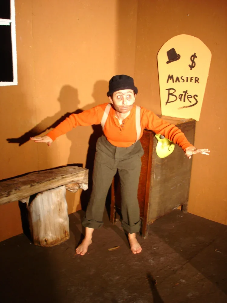 Person in costume standing in a room with a sign that reads 'Master Bates,' wearing an orange shirt, gray pants, a black hat, and suspenders, with wooden furniture and a brown wall in the background.