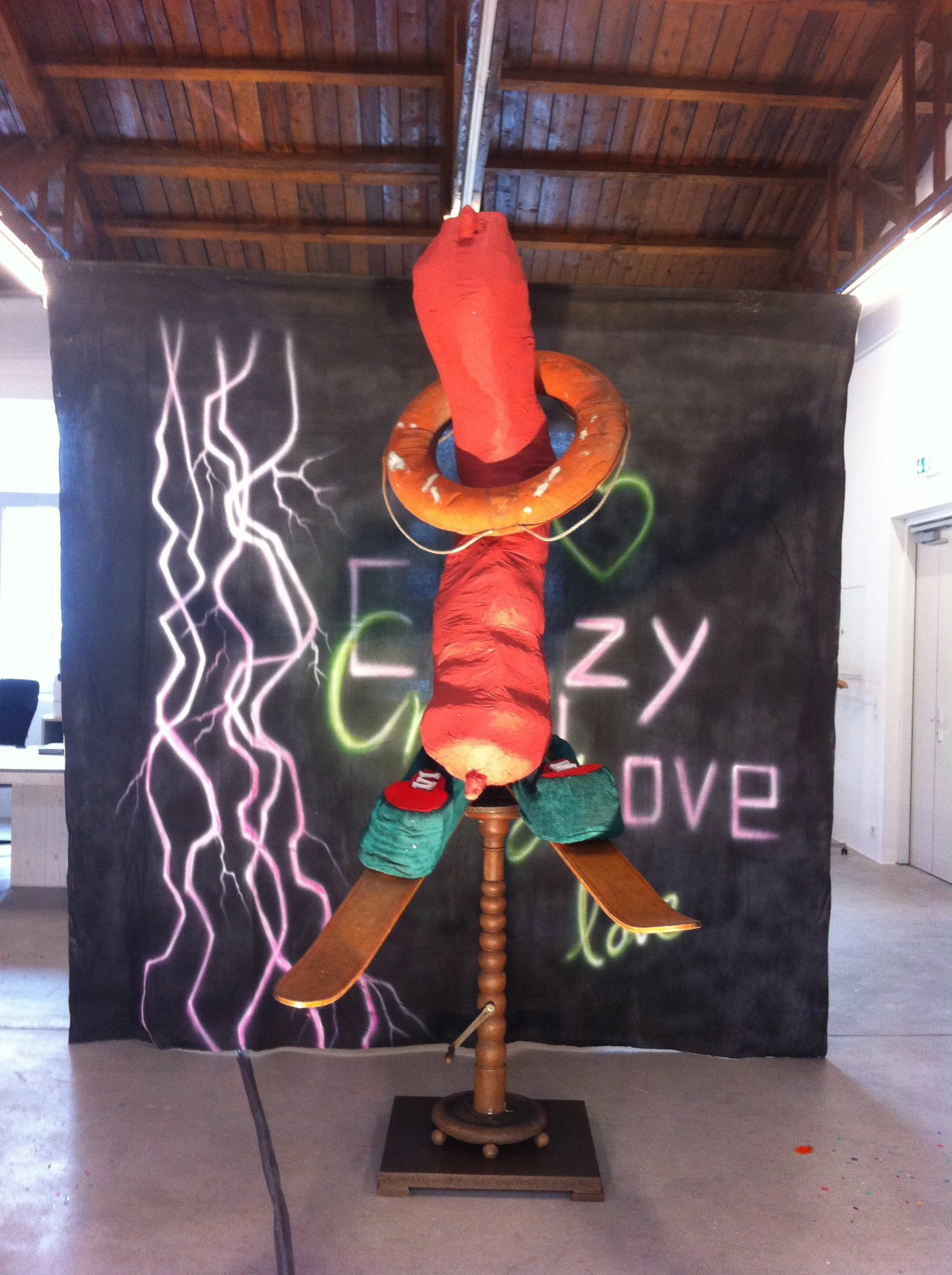 An art installation featuring a hanging puppet dressed like a clown, with colorful clothing, a life buoy around its neck, and propeller blades attached to its body. The background has a dark backdrop with neon-style lightning and the words 'Crazy Love' written in colorful neon lights.