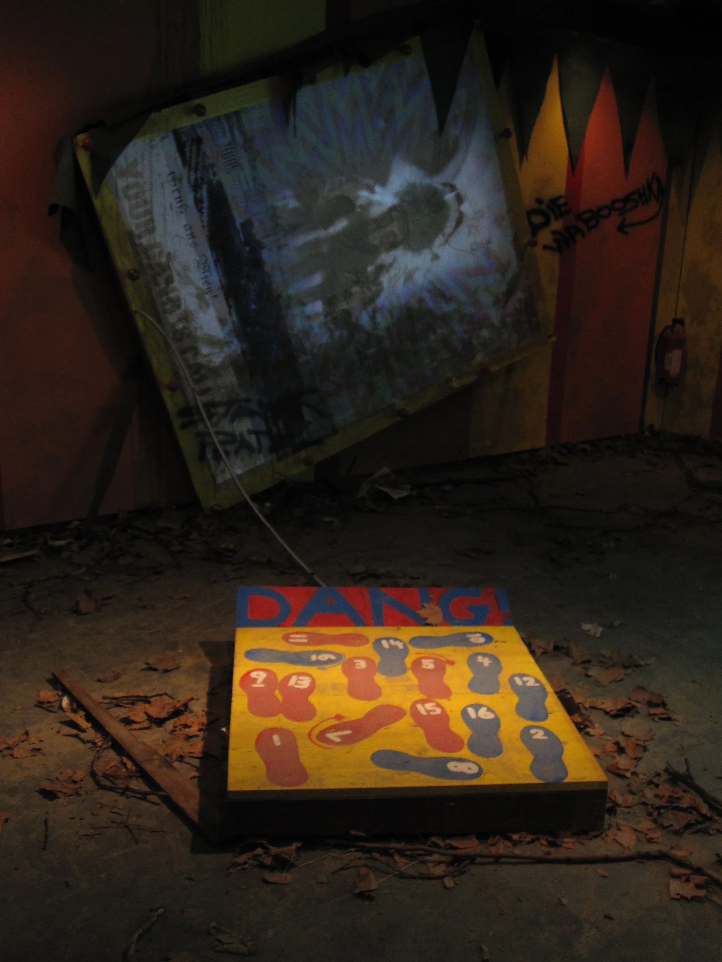 A broken outdoor game with colorful wooden pieces and a large sign that says 'DANG' on the ground, with a fallen billboard in the background that appears to have an artistic image of a dog or wolf.