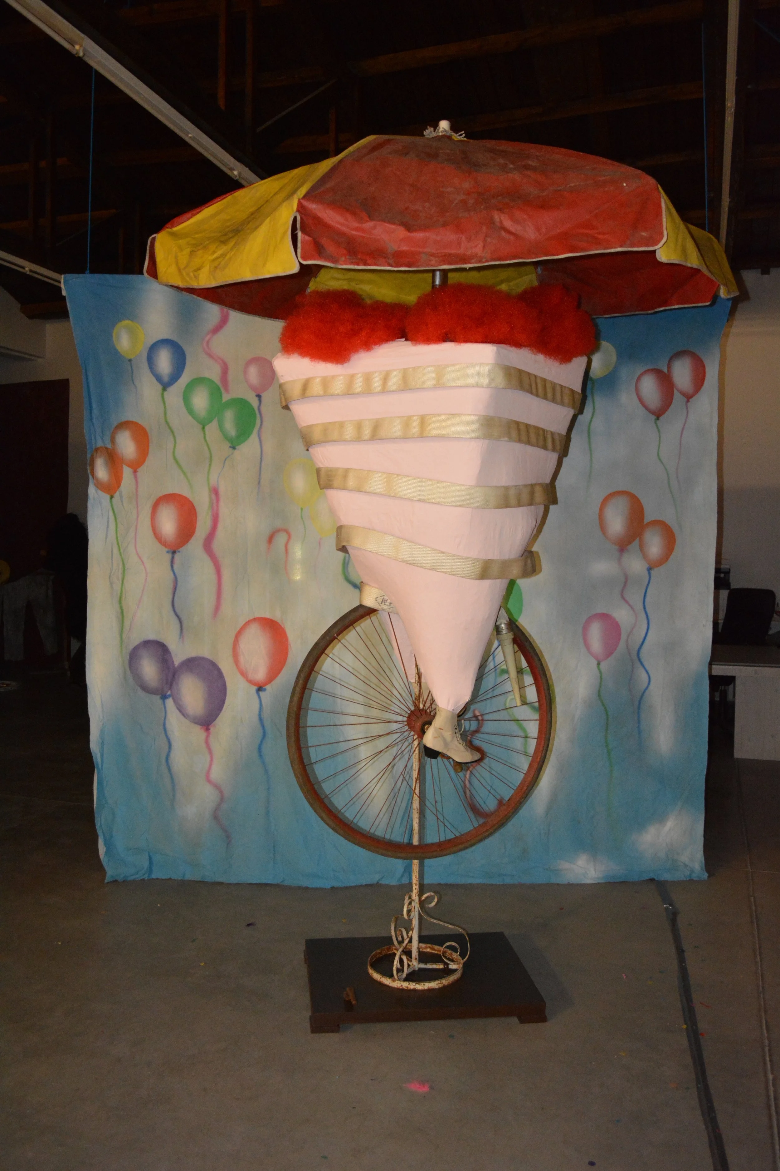 A street performer or art installation features a hybrid bicycle with a large ice cream cone on the front and a multicolored umbrella on top. The background has a colorful balloon-themed curtain.