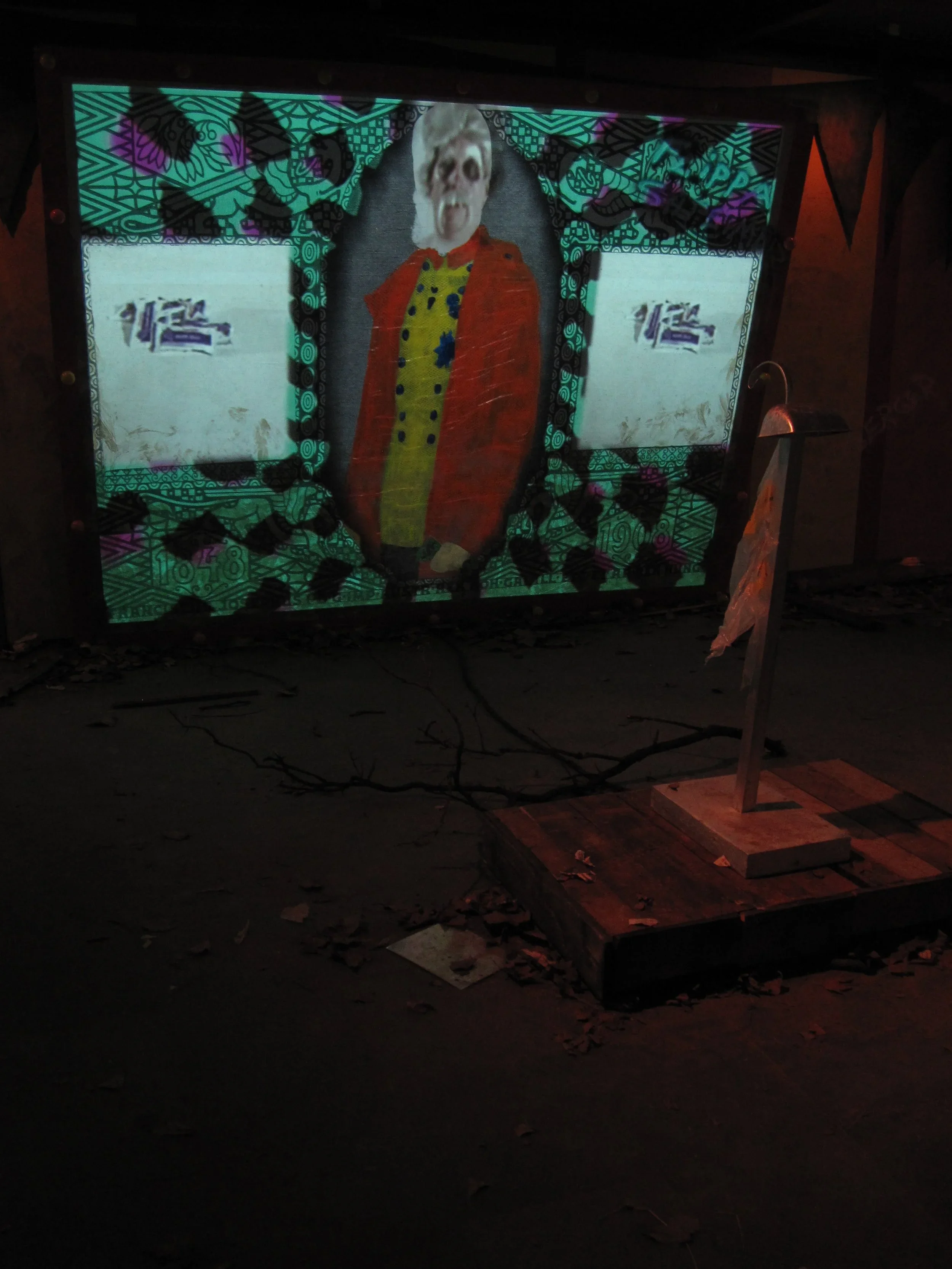 A vertical illuminated art display showing a figure with a painted costume and a white face, surrounded by colorful patterns, with two white screens on either side with the number 12 on them.