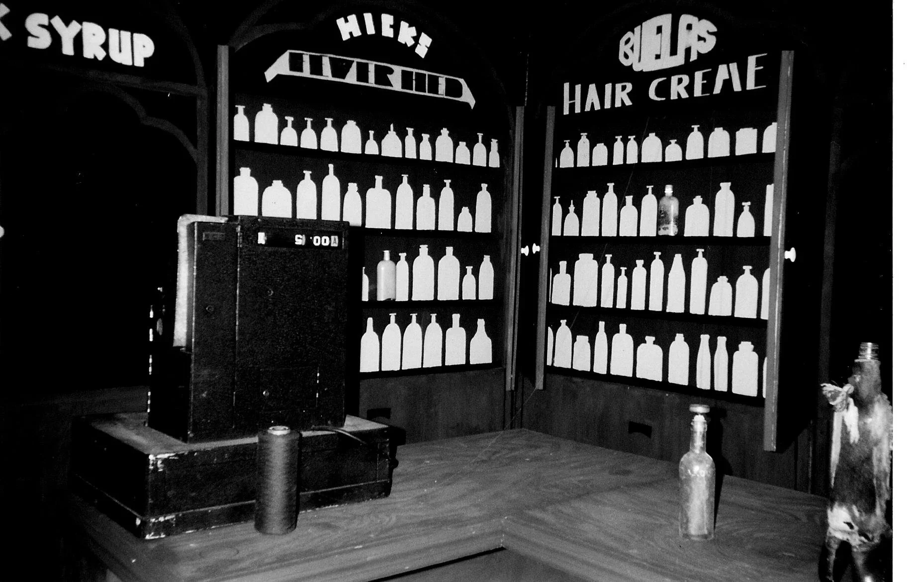 A black and white photo of a bar with shelves filled with bottles, signs mentioning syrup, hair, and nails, and a cash register on the wooden counter.