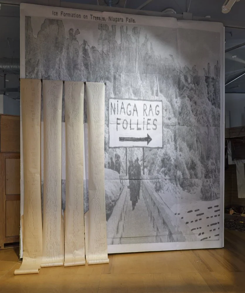 Large mural of a snowy mountainous landscape with a sign pointing right that reads "Niagara RAG FOLLIES." Next to the mural is a set of vertical beige roller shades partially covering it.