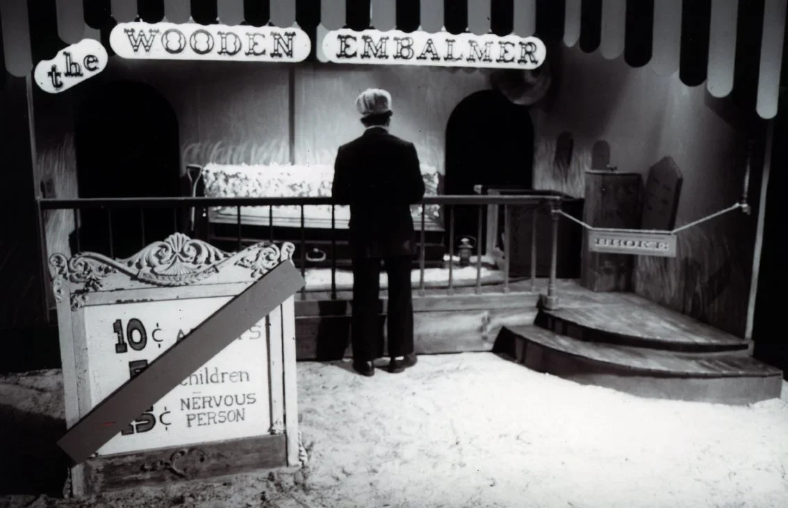 An indoor scene with a person standing in front of a cotton candy stand called 'The Wooden Embalmer' with a sign indicating prices, and a small staircase with a rope barrier. It appears to be a vintage or themed setting.