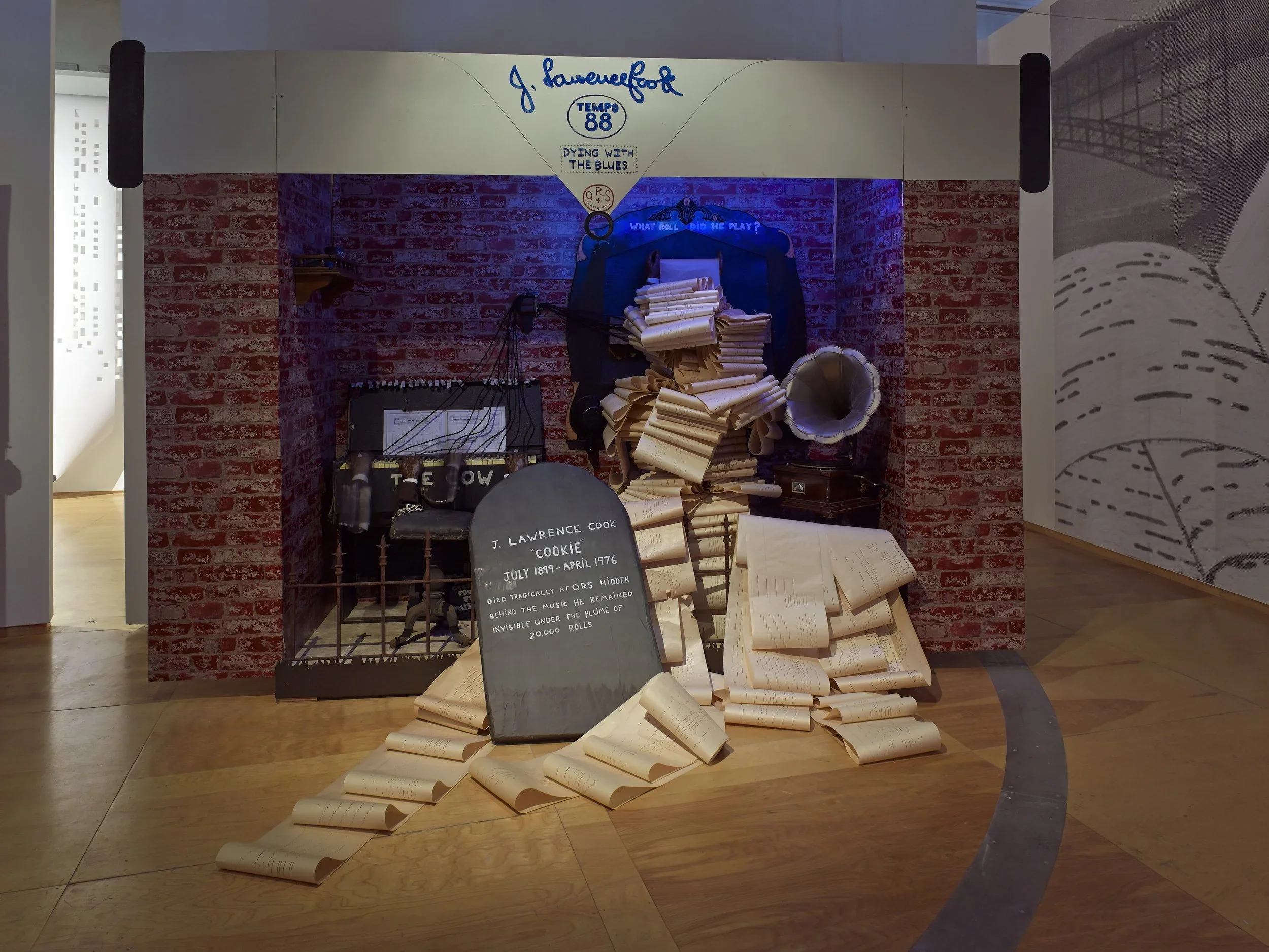 A museum display dedicated to J. Lawrence Cook, featuring a large pile of scrolls and papers, a sign with his biography, an old radio, a vintage phonograph, and a photo backdrop with a brick wall and musical notes.