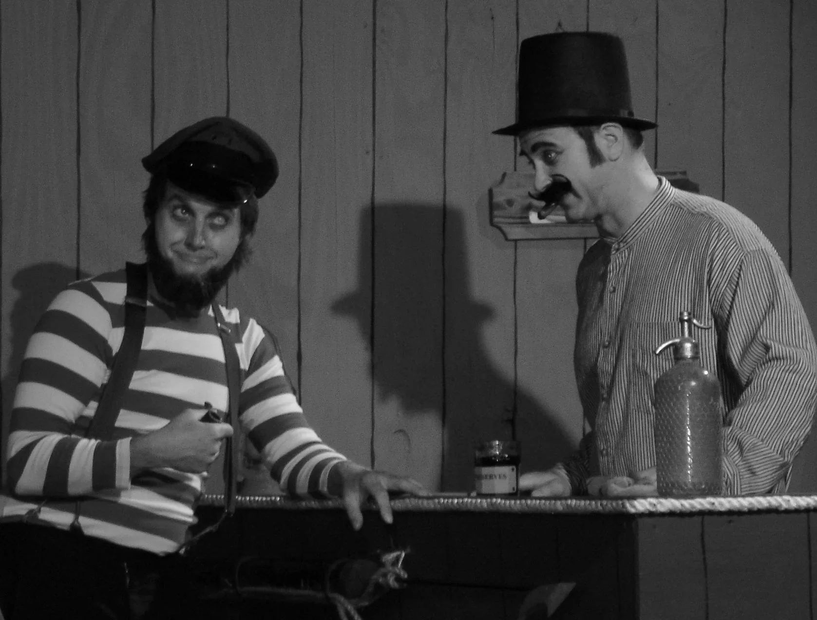 Two men dressed as comic or clown characters standing at a table with a wood-paneled wall in the background. One man has a beard and is wearing a striped shirt and suspenders, and the other man has a mustache, a tall hat, and face makeup.