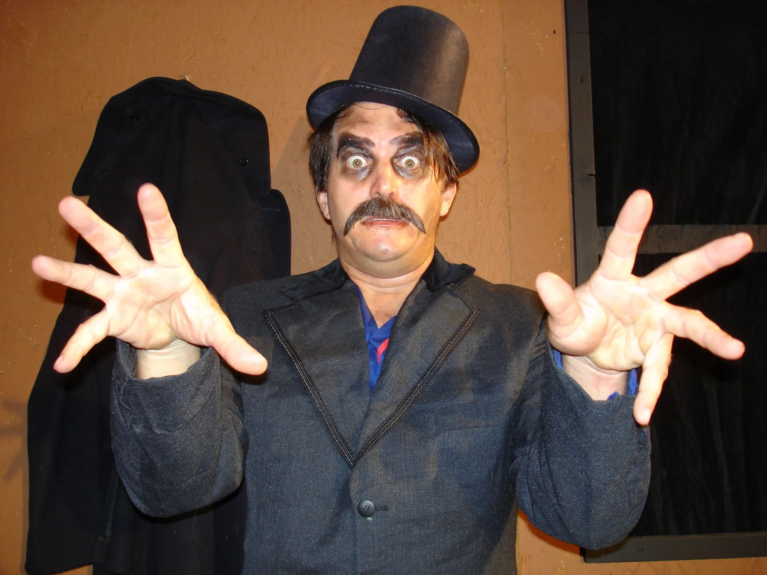 A man dressed in a costume with a top hat, dark coat, and theatrical makeup, making a spooky gesture with his hands.