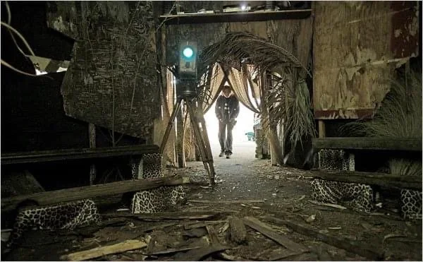 Person walking through a makeshift tropical hut entrance with jungle foliage, rough wooden benches, and debris on the ground.