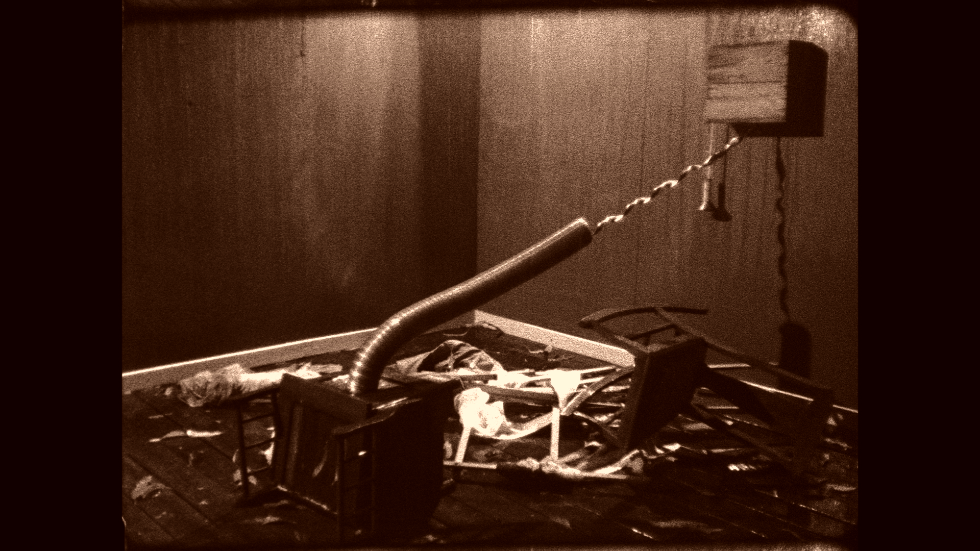 A burned and damaged metal bed frame with a twisted headboard and scattered debris, next to an electrical outlet on a burnt wall.