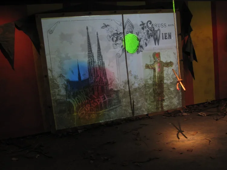 A projection on a wall showing images of the Vienna Cathedral, graffiti, and a large person in traditional clothing, with the text 'Gruss aus Wien' and a green spiral pattern in the center.