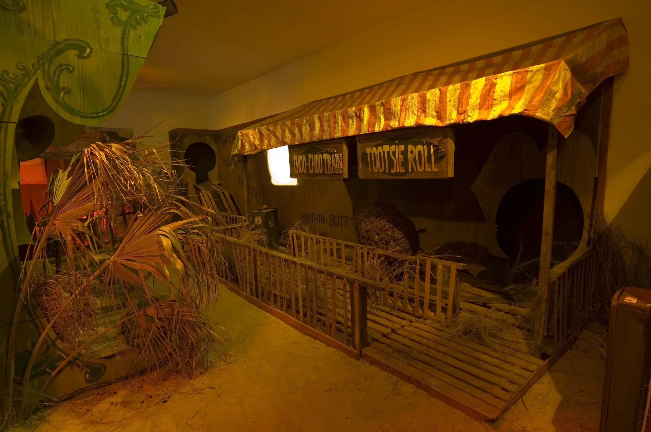 Indoor scene with a small wooden fenced area and signs for snacks like "Tootsie Roll" and "Loco-Choco Treat." Artificial plants and a colorful striped canopy over the fence are also visible.