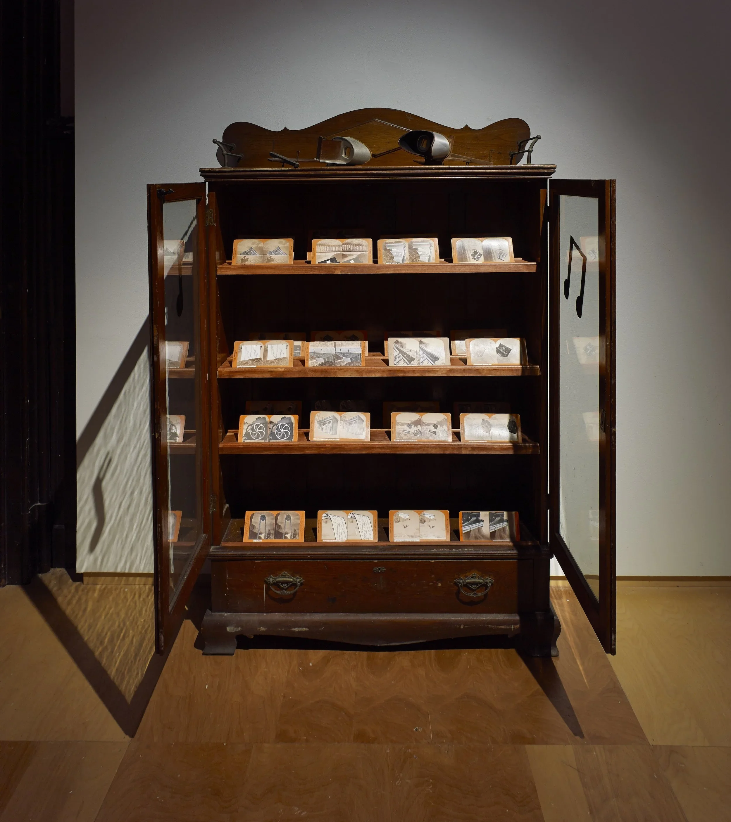 A vintage wooden cabinet with glass doors, containing small display cards and musical note symbols, with a stereo on top.