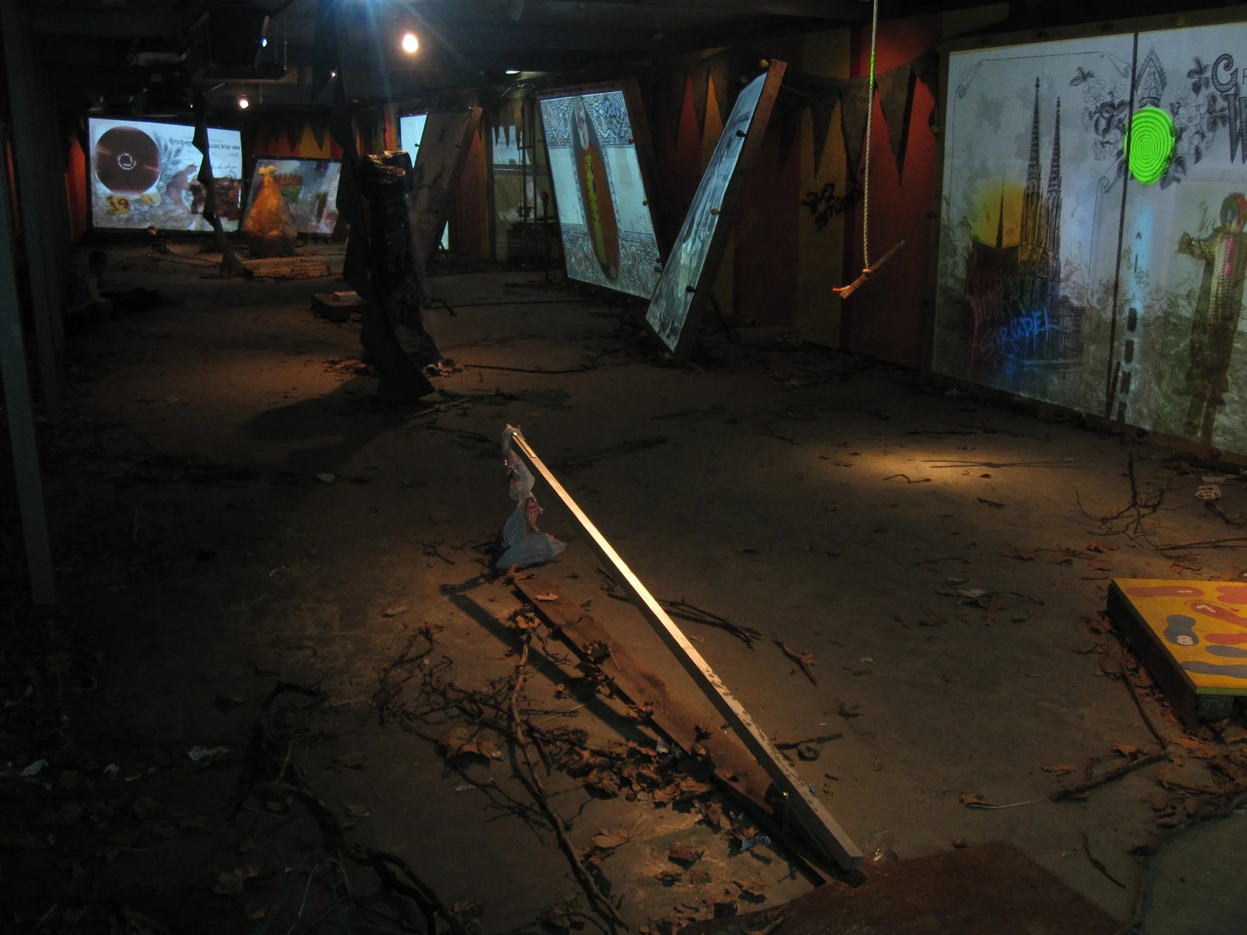 Dark indoor space with damaged sign, debris, fallen branches, and windows covered with graffiti or artwork.
