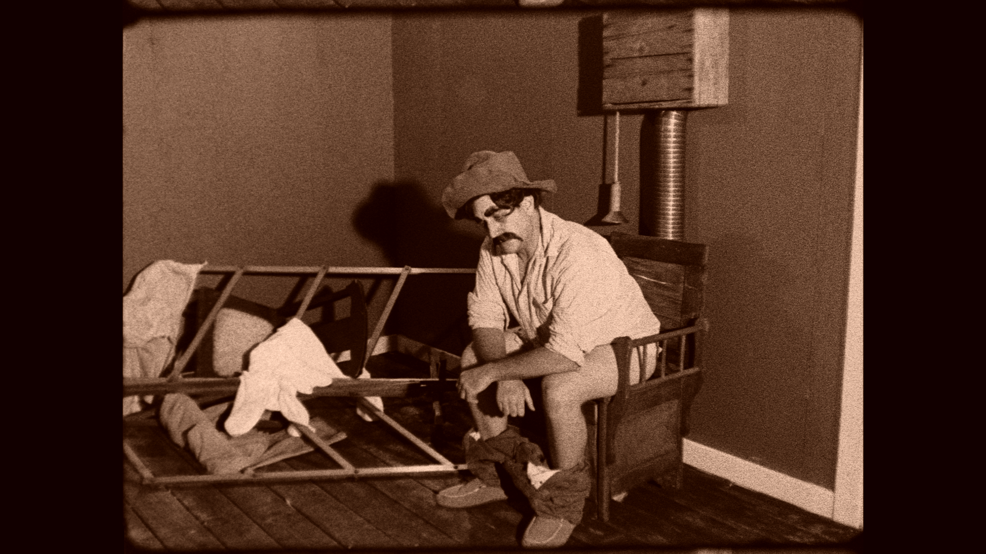 A person with a mustache, sunglasses, and a hat, sitting on the edge of a wooden bed frame, appearing to be a mime or performer in costume. The bed's metal frame is partially disassembled with some clothes draped over it. The room has wooden flooring and a plain wall with a pipe and wooden block attached to the wall behind.