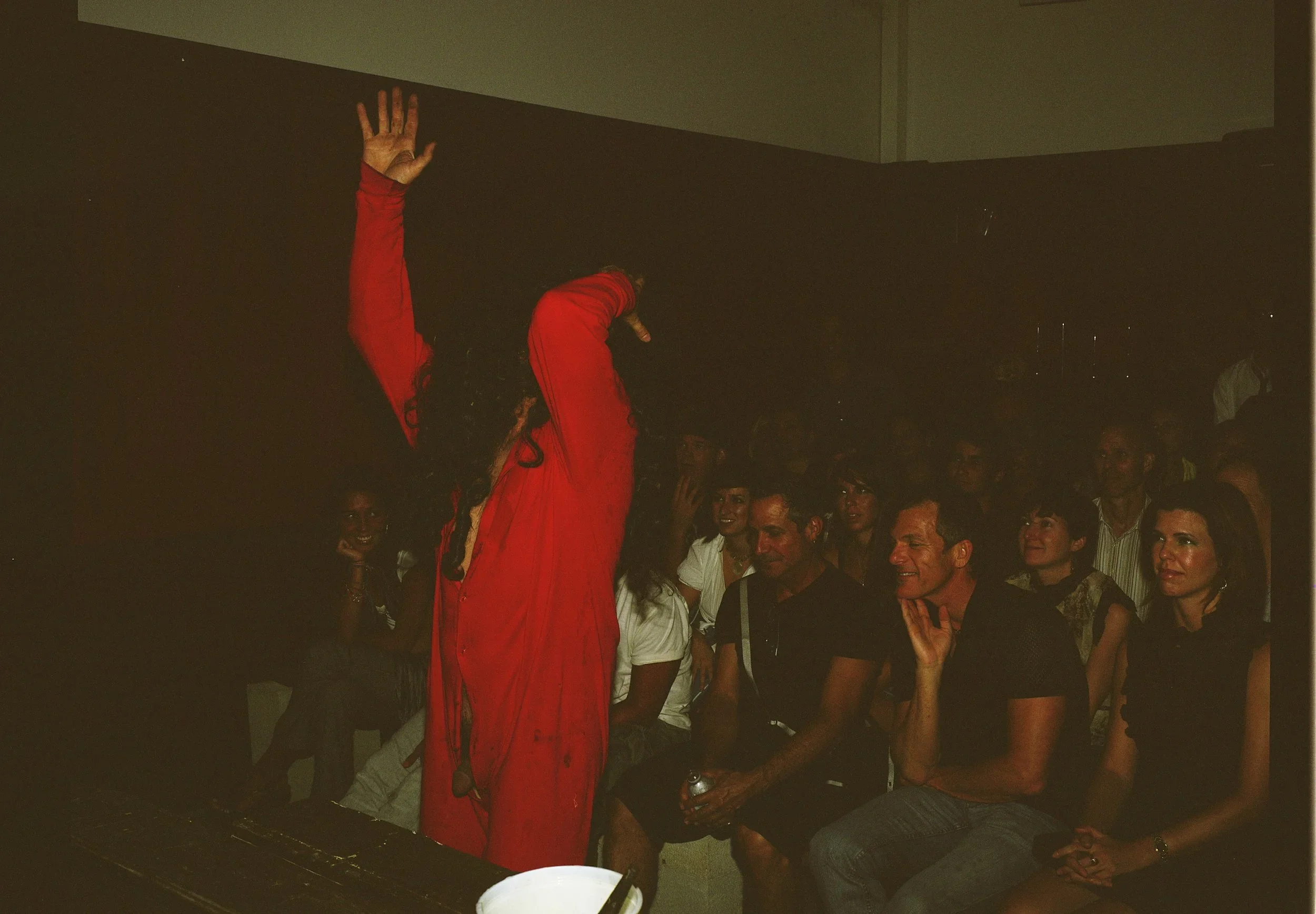 A woman in a red dress is performing on stage in front of an audience. The audience members, including several men and women, are seated and watching her performance.
